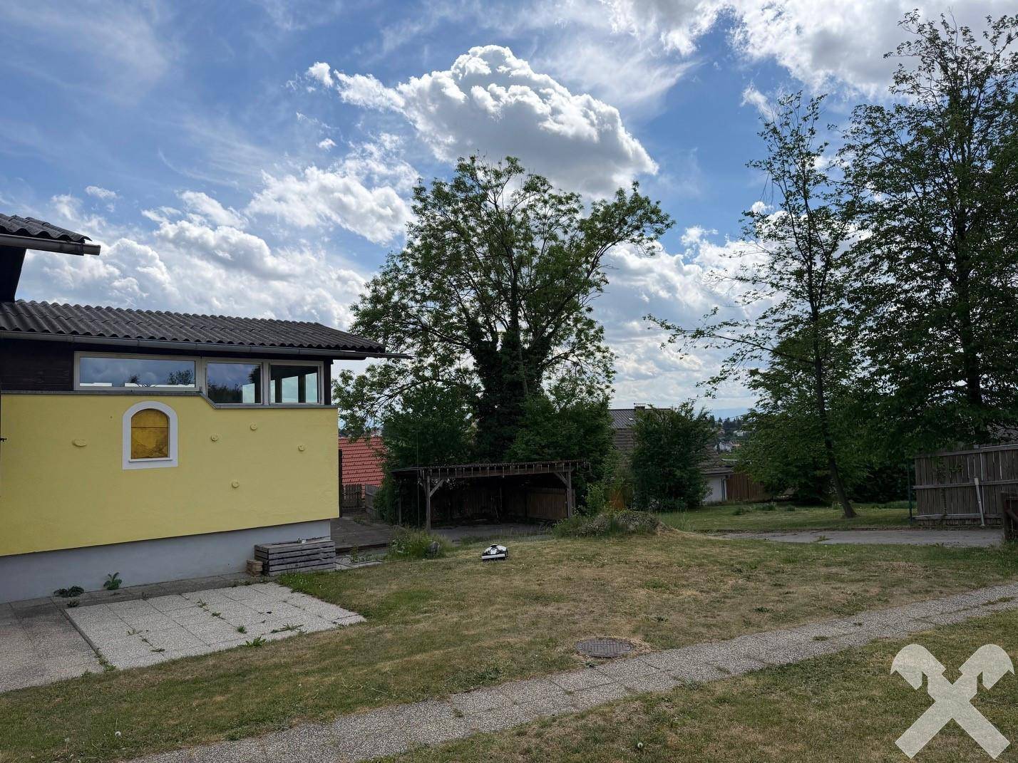 Seitenansicht des gelben Hauses mit Rasenfläche und einer Holzkonstruktion im Hintergrund unter bewölktem Himmel.