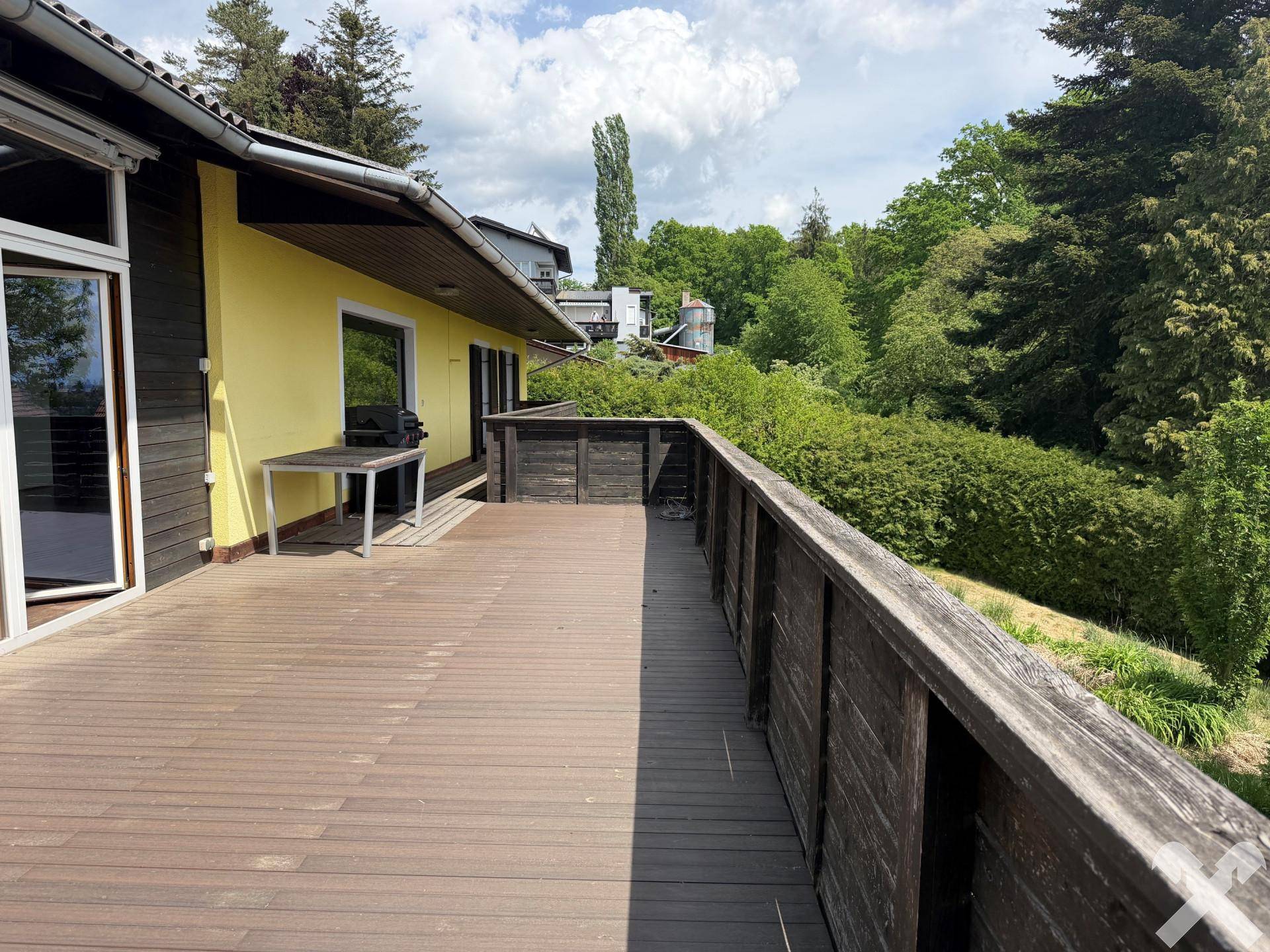 Langer Holzbalkon entlang der Hausfassade mit einem kleinen Tisch und Blick ins Grüne.