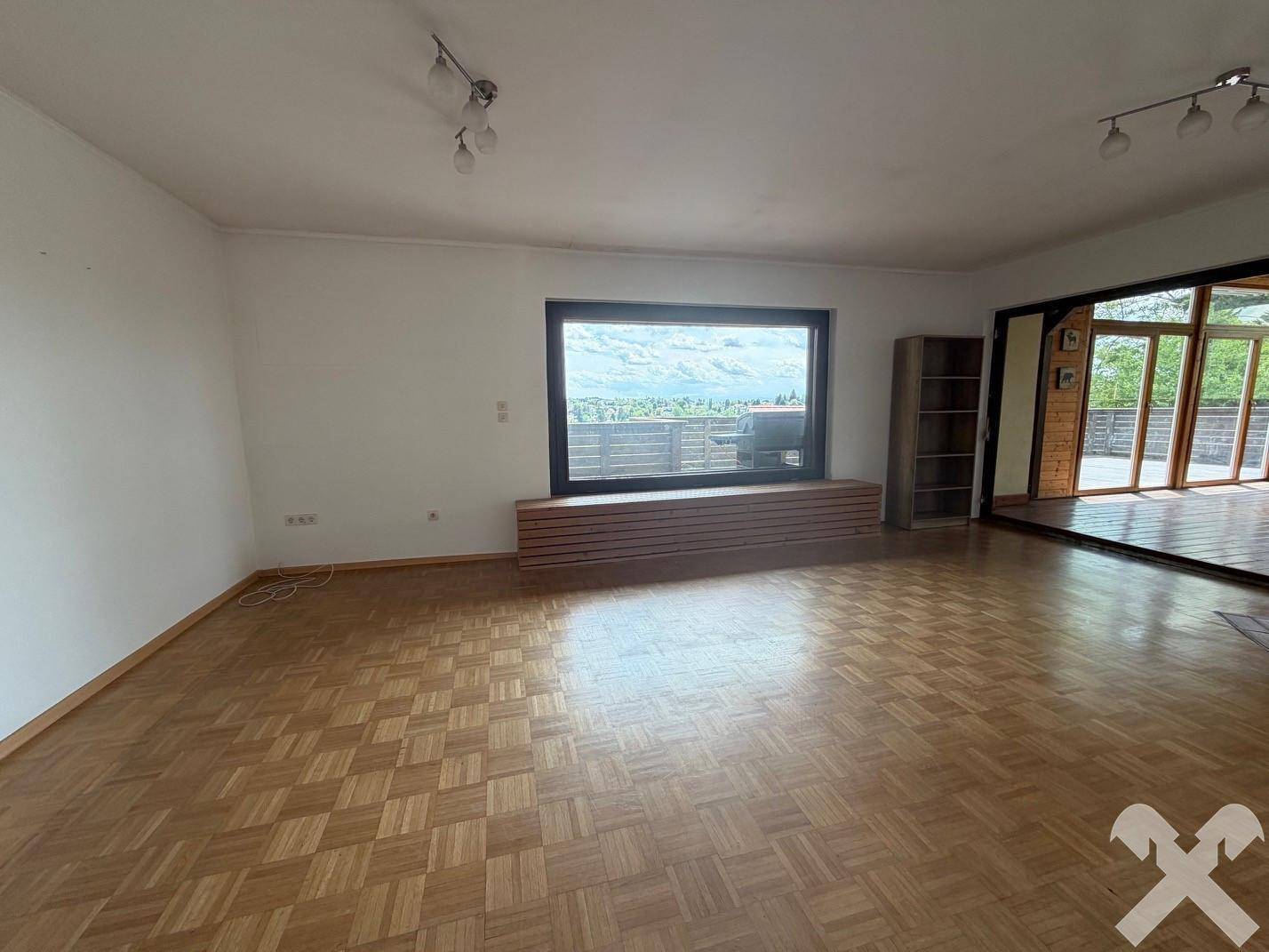 Geräumiges Wohnzimmer mit Parkettboden, weißen Wänden und einem großen Fenster mit Blick ins Freie.