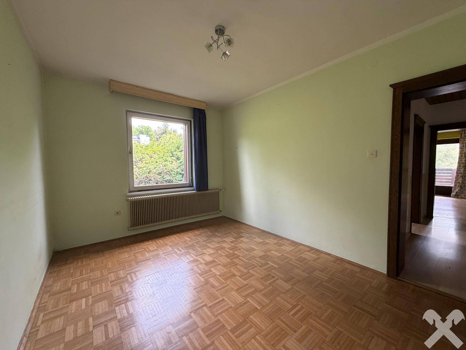 Helles Zimmer mit hellgrünen Wänden, Parkettboden und einem Fenster mit Blick ins Grüne.