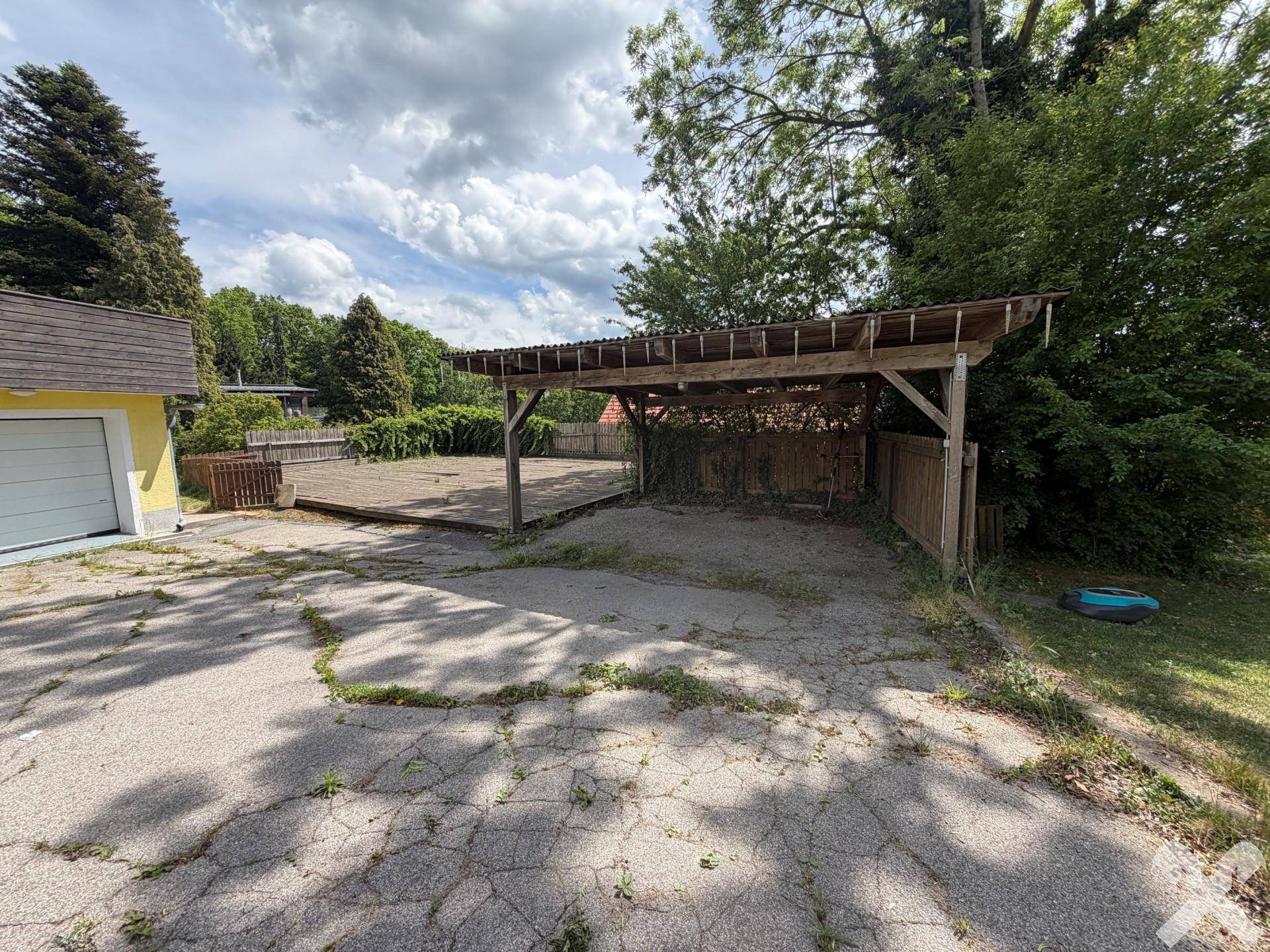Außenbereich mit Carport, Holzterrasse und asphaltierter Zufahrt.