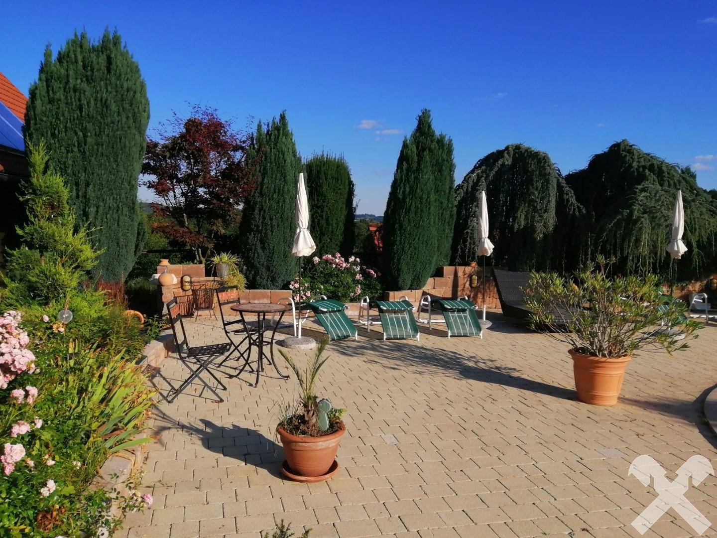 Weitläufiger Gartenbereich mit gepflasterter Fläche, Liegestühlen und vielfältiger Bepflanzung.