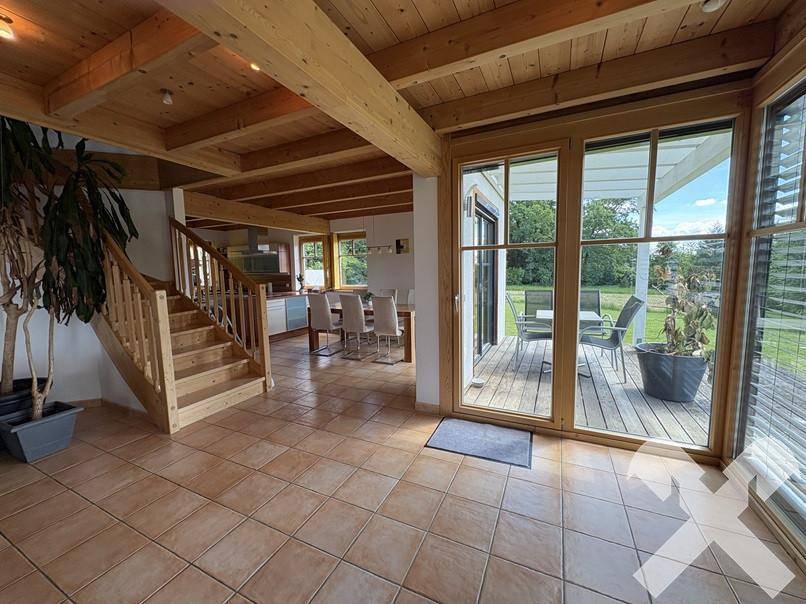 Offener Wohn- und Essbereich mit Fliesenboden, Holztreppe und großen Fenstern zur Terrasse.