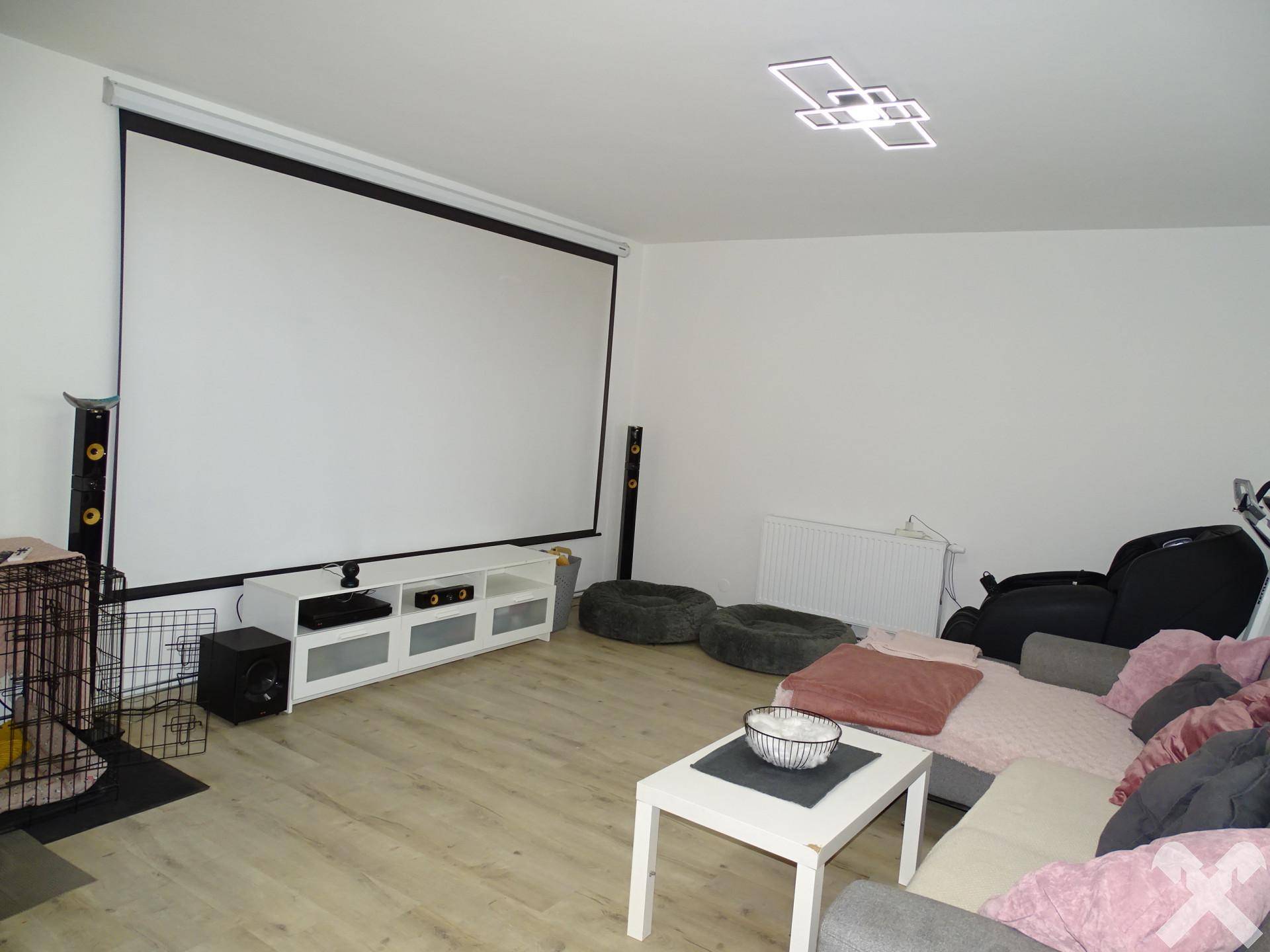 Geräumiges Wohnzimmer mit Projektionsleinwand, hellem Laminatboden und moderner Deckenleuchte.