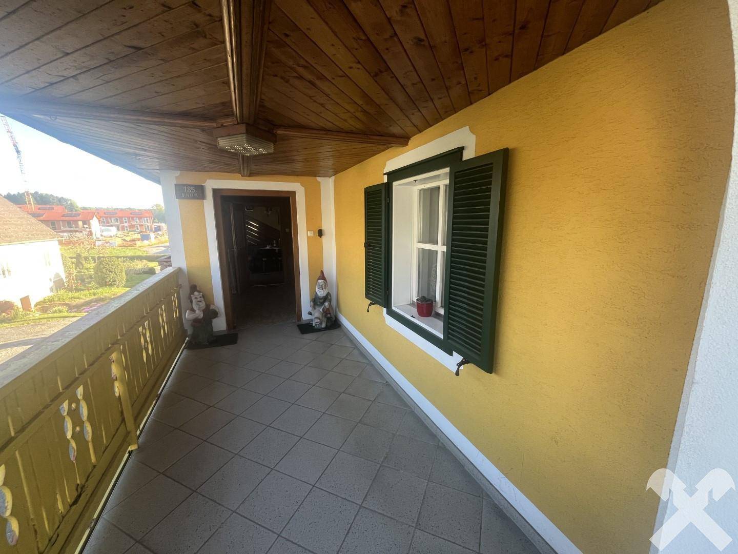 Überdachter Eingangsbereich mit Holzbalkendecke, Fliesenboden und einem Fenster mit grünen Fensterläden.