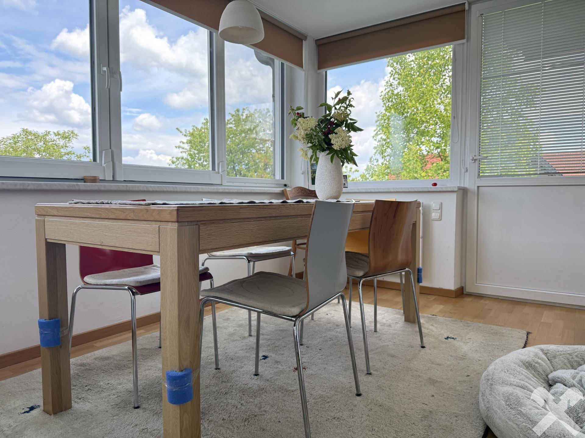 Heller Essbereich mit Holztisch und Stühlen, umgeben von großen Fenstern mit Blick ins Grüne.