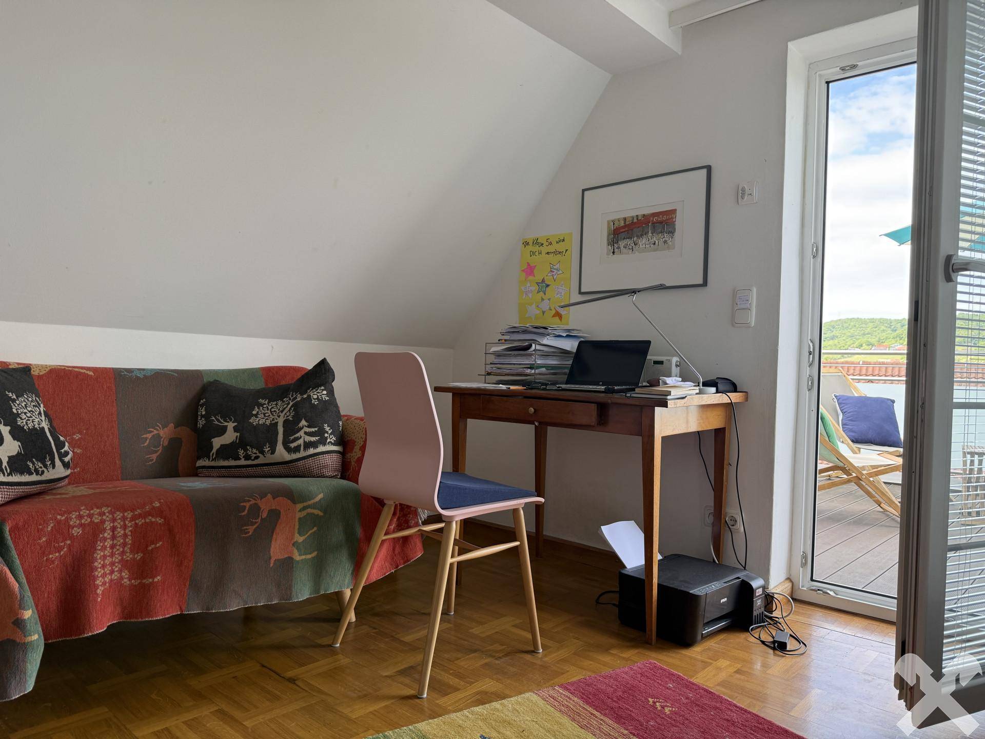 Arbeitsbereich mit Sofa, Schreibtisch und Zugang zu einer Terrasse, unter einem Dachschrägenfenster.