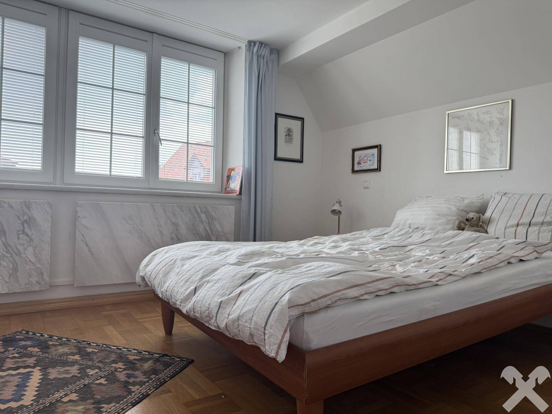 Helles Schlafzimmer mit Holzboden, Doppelbett und Fenstern mit Jalousien für Privatsphäre.