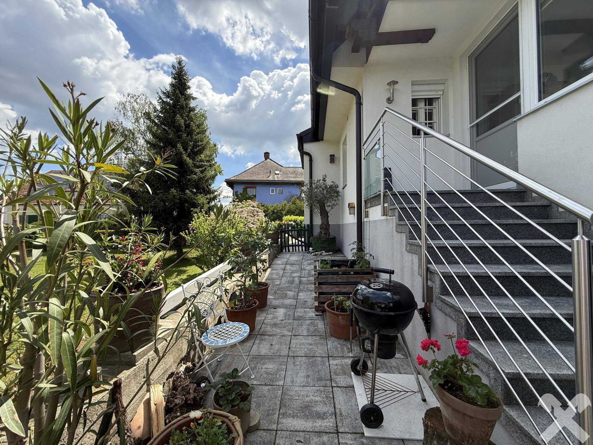 Gepflasterter Seitenweg mit vielen Topfpflanzen, Grillbereich und Treppe zum oberen Bereich.