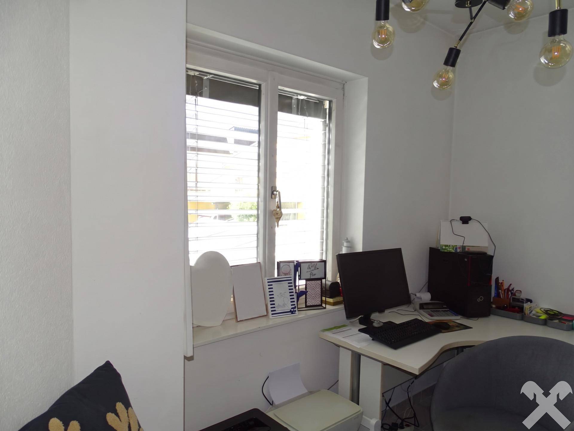 Heller Arbeitsbereich mit Schreibtisch am Fenster und moderner Beleuchtung.