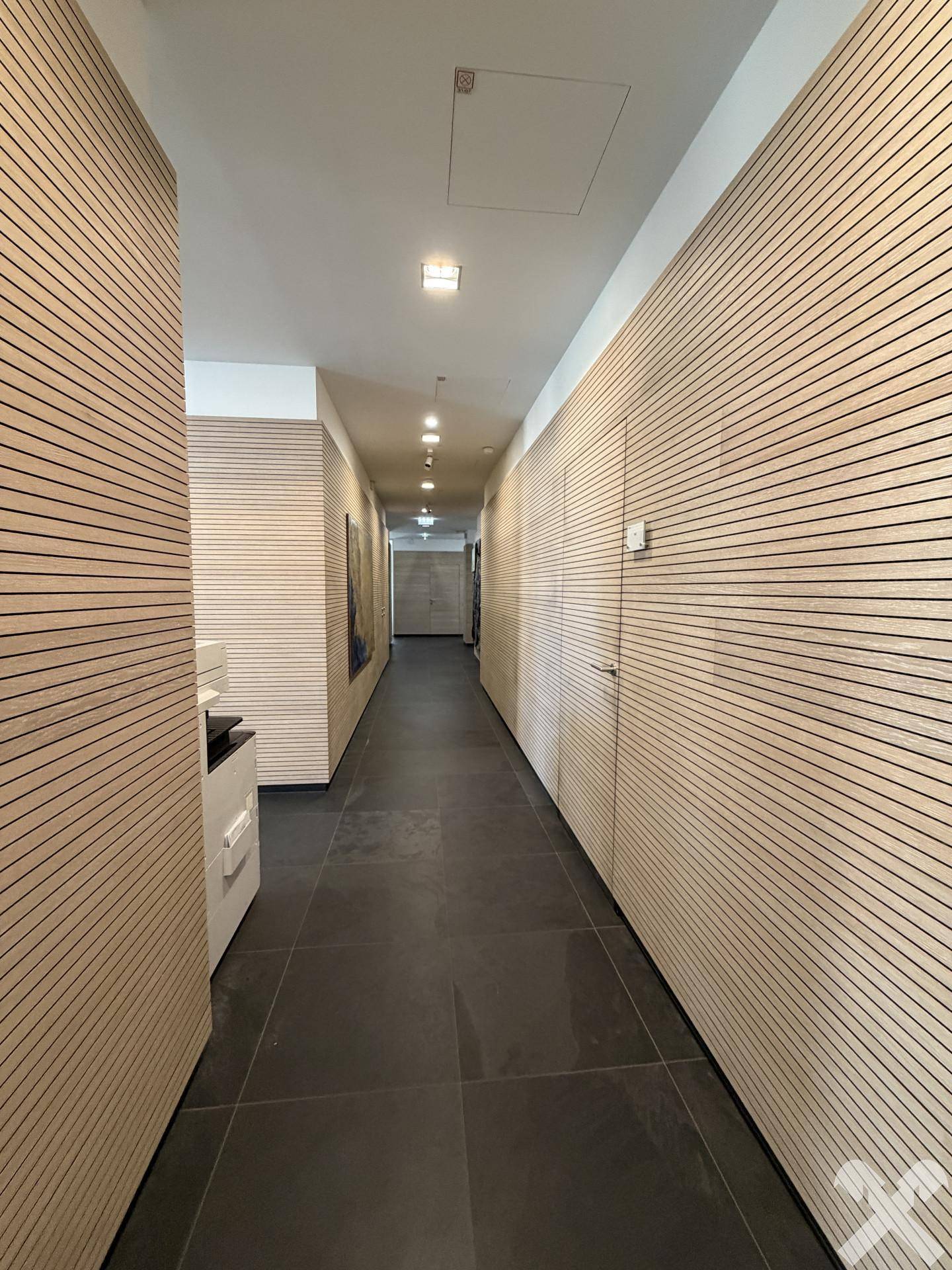 Langer Flur mit Holzlamellenwänden, dunklem Fliesenboden und Deckenleuchten.