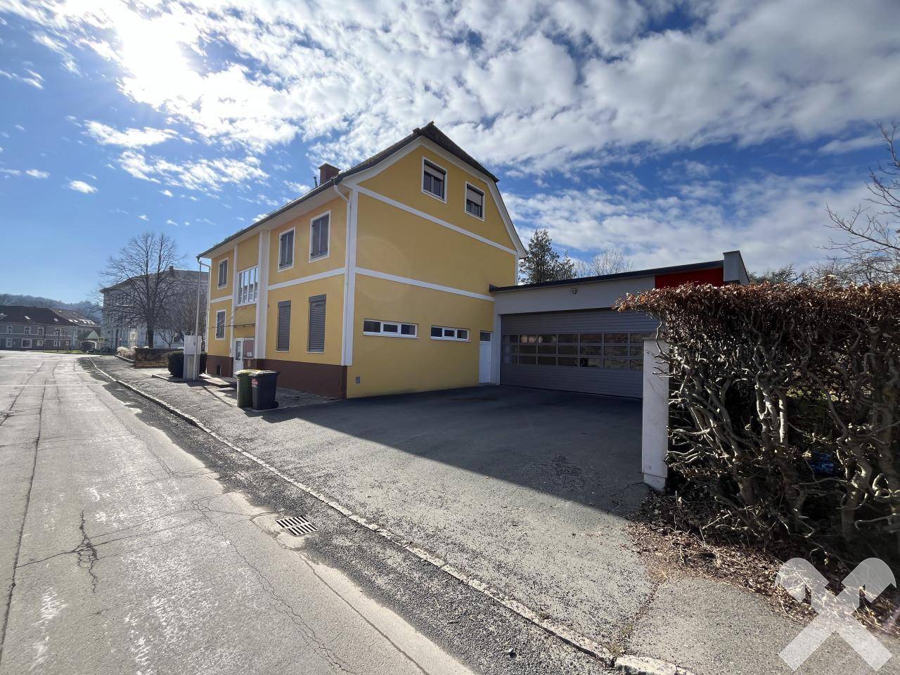 Gelbes Gebäude mit Garage und Straße davor, umgeben von Bäumen und Hecken.