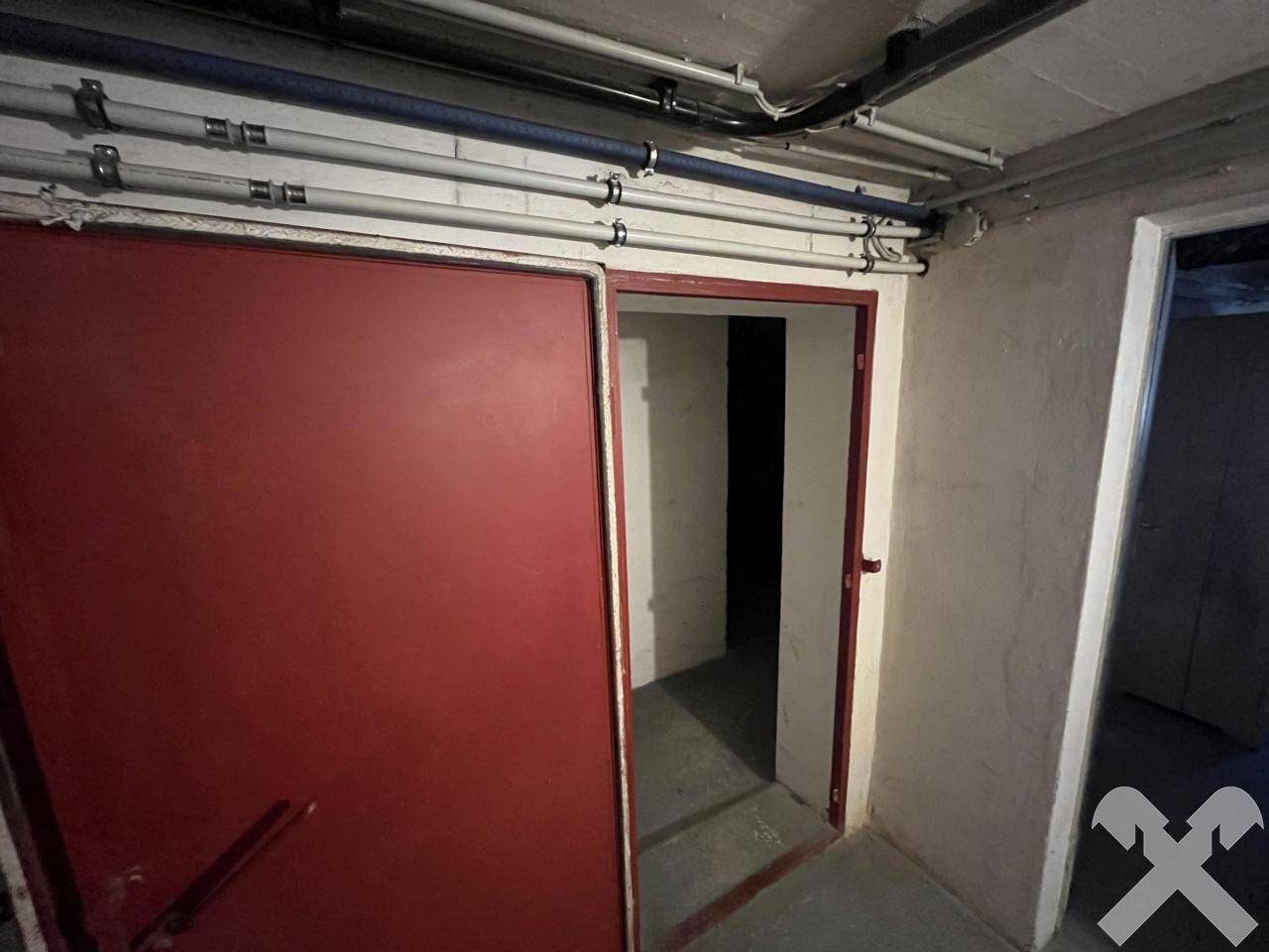 Kellerbereich mit einer großen roten Tür und sichtbaren Deckenrohren, führt zu weiteren Räumen.