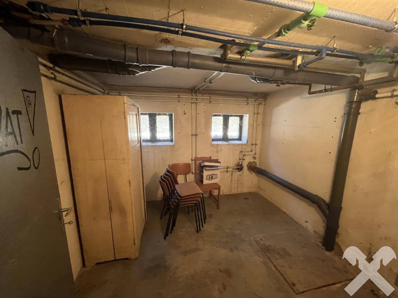 Kellerraum mit sichtbaren Rohren an der Decke, kleinen Fenstern und einem Schrank für Lagerzwecke.
