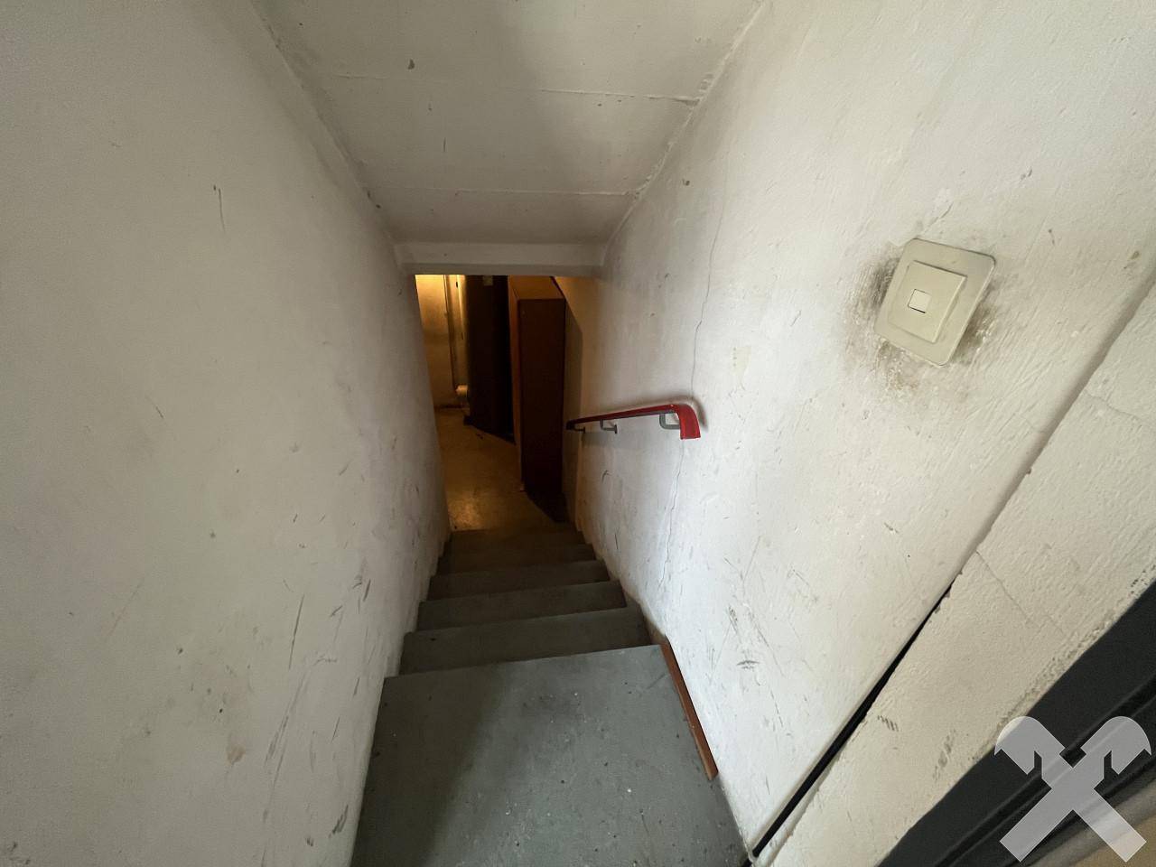 Treppenabgang in den Keller mit schlichten, weiß gestrichenen Wänden und einem roten Handlauf für Sicherheit.