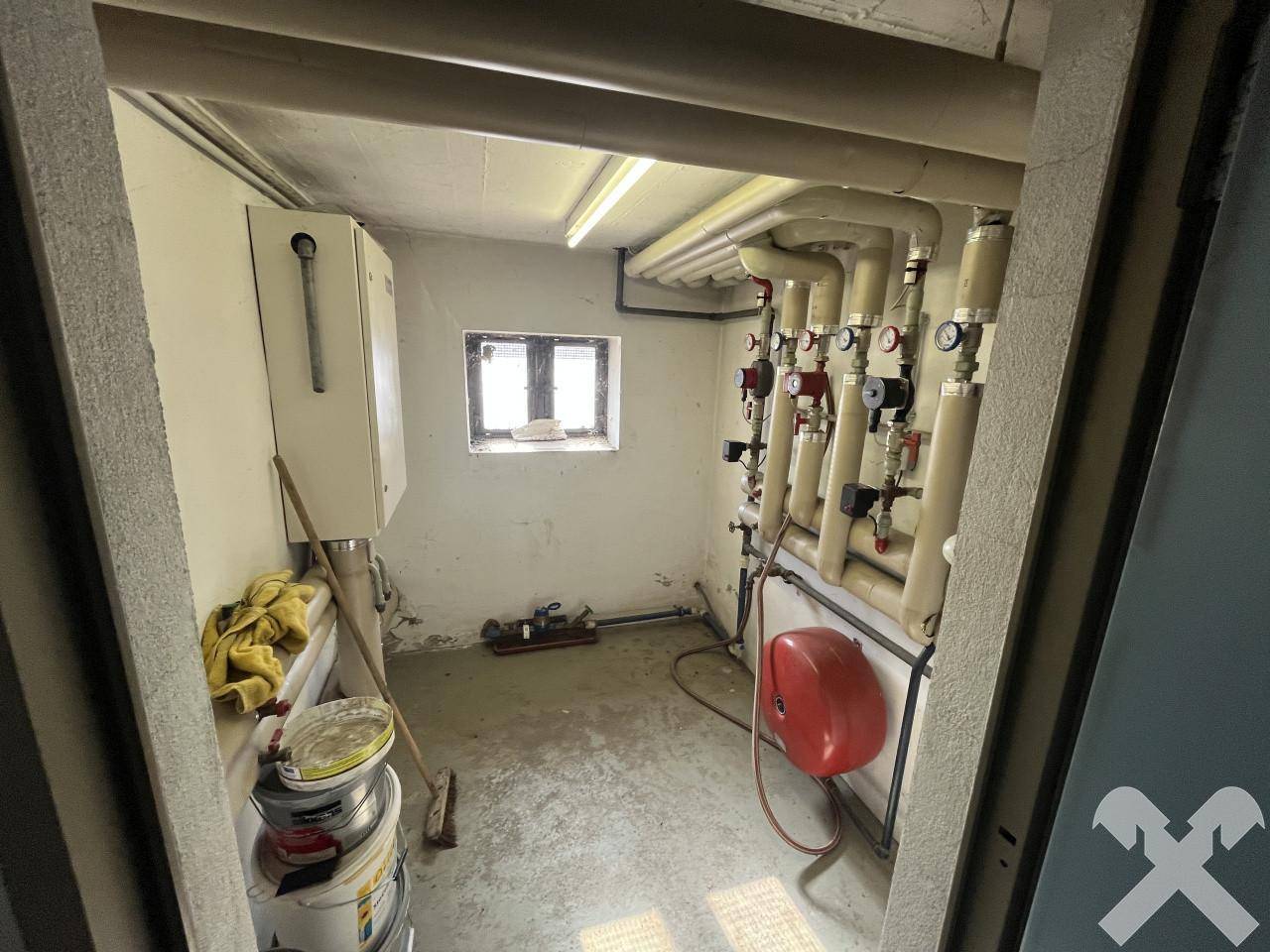 Technikraum mit Heizungsanlage, zahlreichen Rohren und einem kleinen Fenster, dient als Versorgungszentrale.