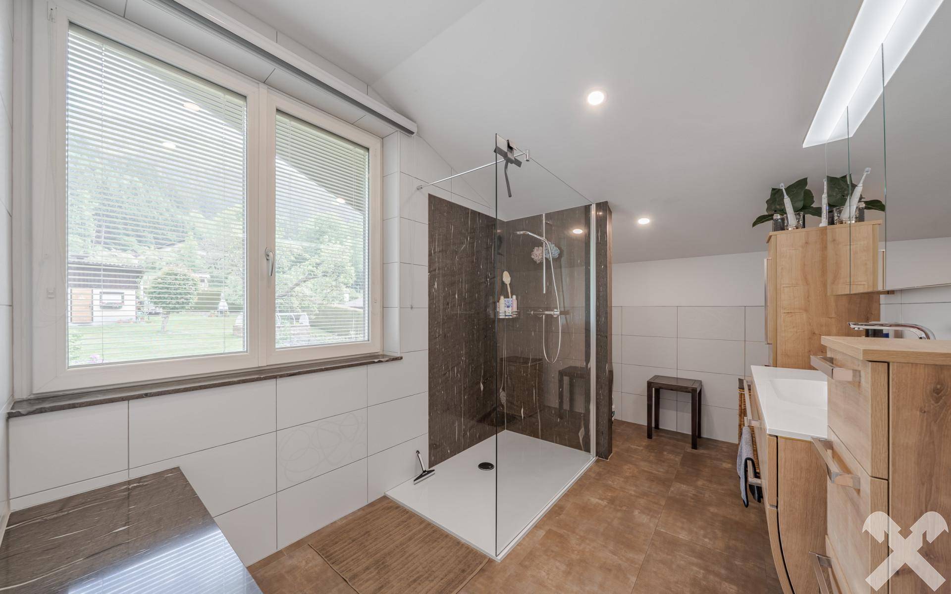 Geräumiges Badezimmer mit großer begehbarer Dusche, Holzwaschtisch und Fenster mit Blick ins Grüne.