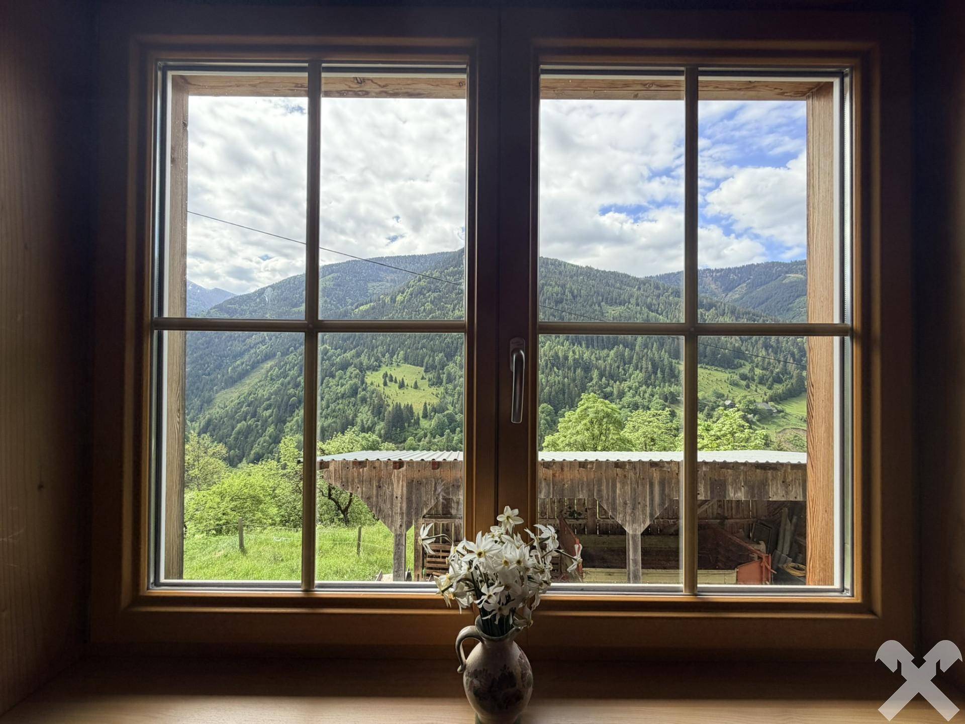Malerischer Ausblick aus einem Holzfenster auf die grüne Berglandschaft und ein traditionelles Nebengebäude.