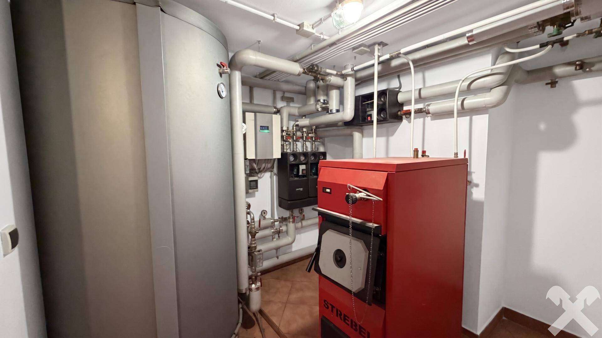 Heizungsraum mit großem Warmwasserspeicher, rotem Heizkessel und sichtbaren Rohrleitungen.