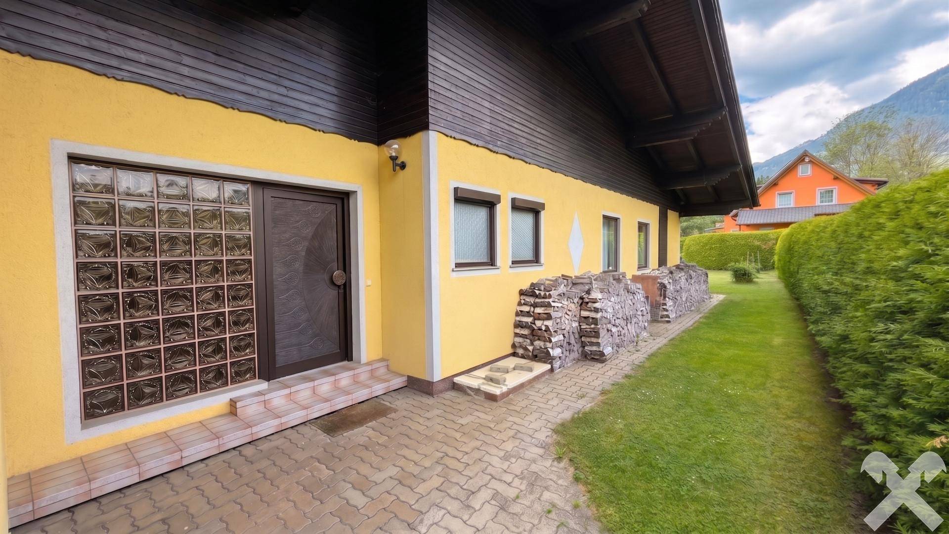 Hausfassade mit Eingangsbereich, Glasbausteinwand, gestapeltem Brennholz und gepflastertem Weg.