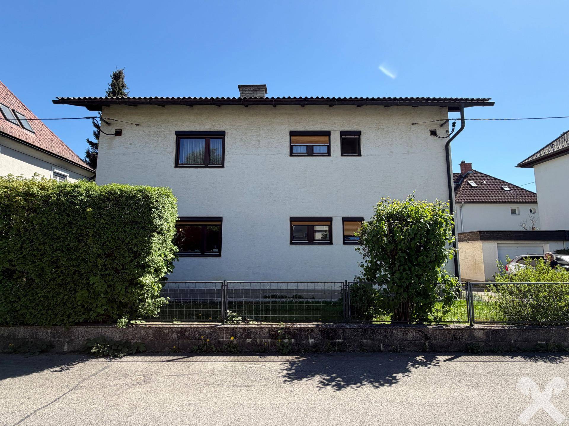 Vorderansicht des Einfamilienhauses mit weißer Fassade, dunklen Fenstern und grüner Hecke.