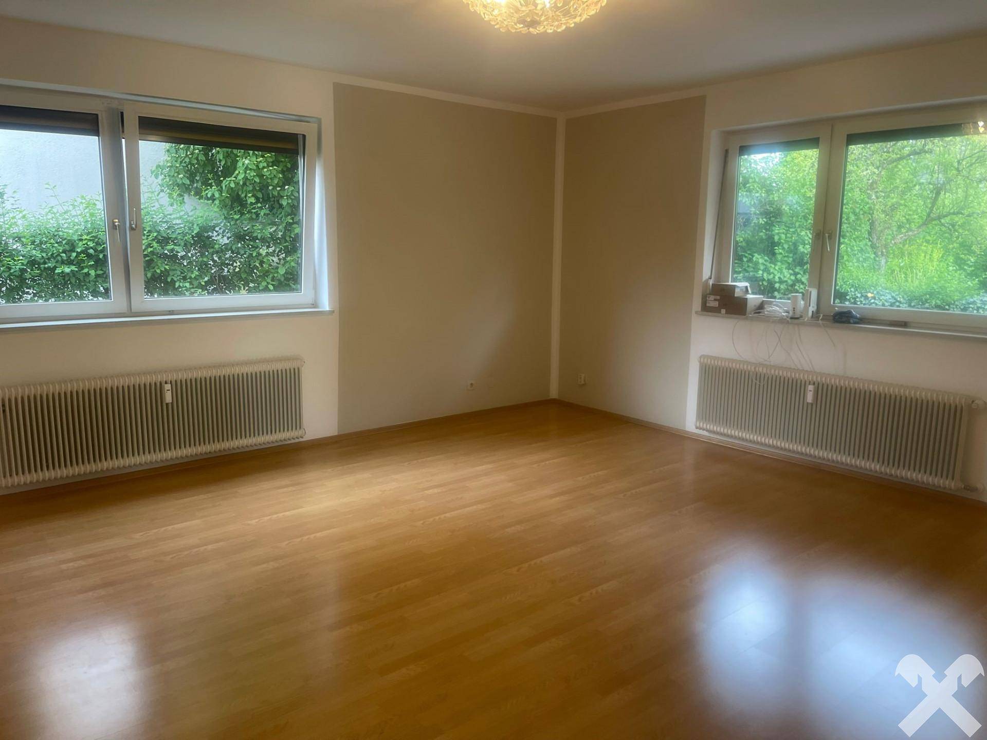 Geräumiges Zimmer mit hellem Parkettboden und zwei großen Fenstern mit Blick ins Grüne.