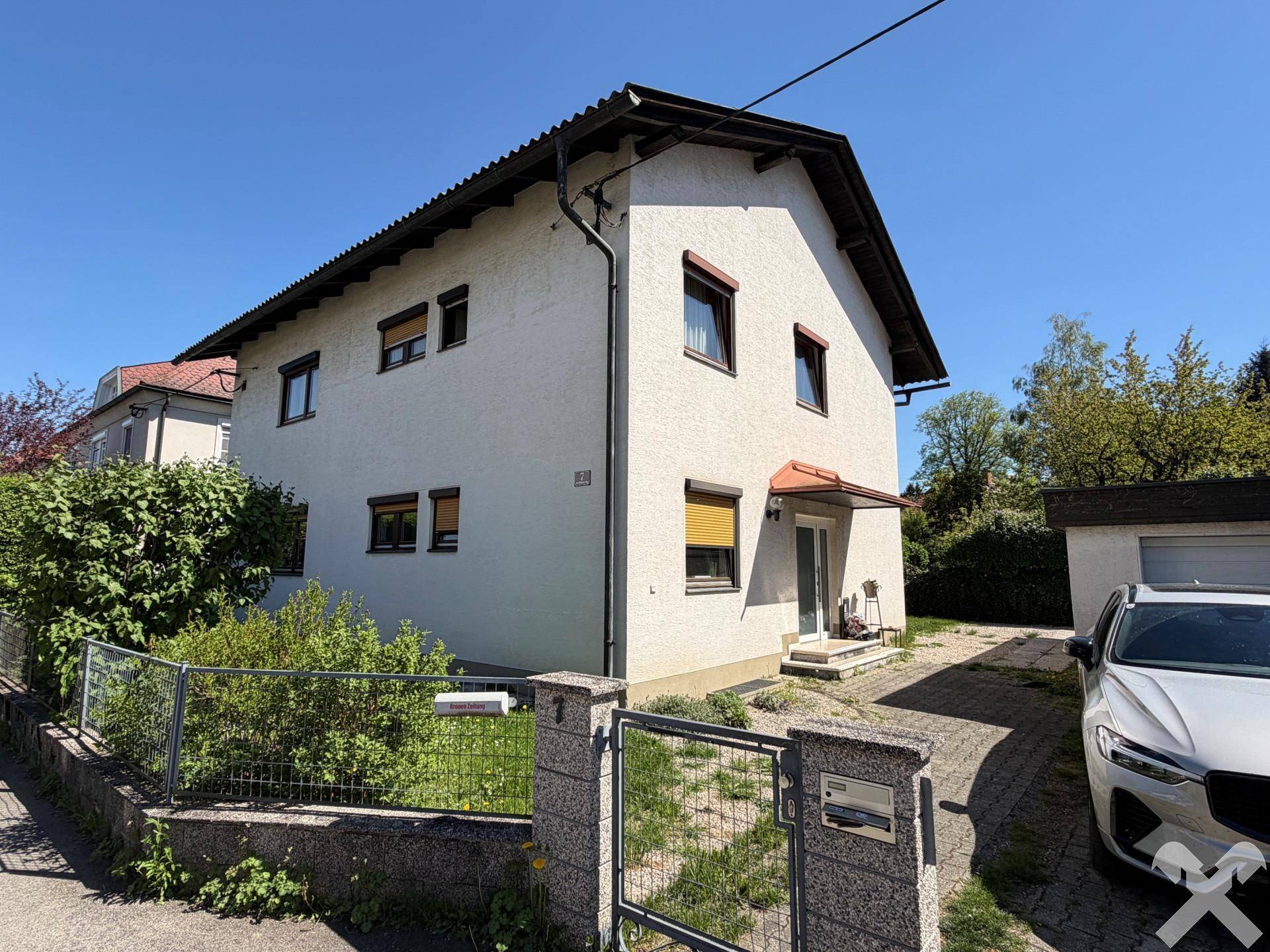 Weißes Einfamilienhaus mit Vorgarten, Garagenzufahrt und geparktem Auto an einem sonnigen Tag.