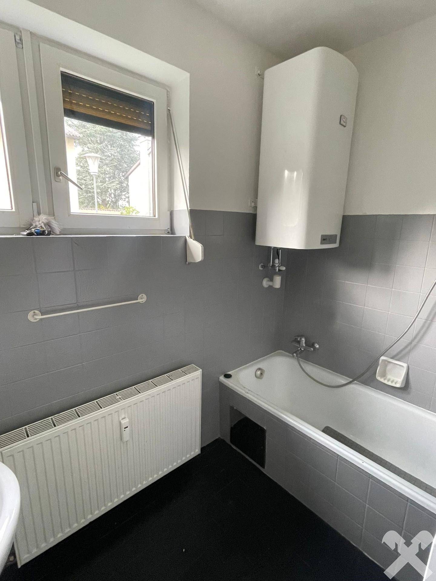 Badezimmer mit Badewanne, Fenster und Wandboiler, gefliest in Grau und Weiß.