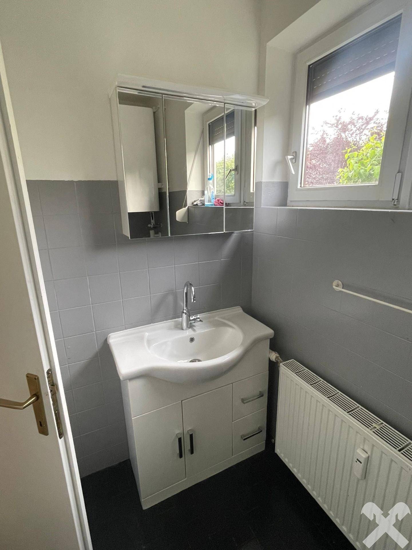 Badezimmer mit Waschbecken, Spiegelschrank und Fenster, teilweise gefliest in Grau.