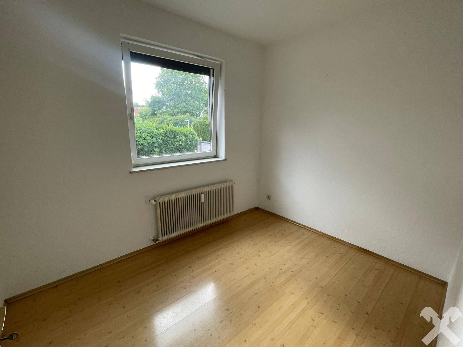 Helles, leeres Zimmer mit Parkettboden und einem Fenster mit Blick ins Grüne.