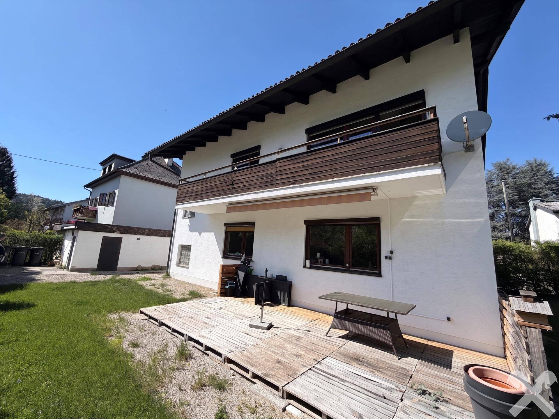 Rückseite des Hauses mit großer Holzterrasse, Balkon und Rasenfläche an einem sonnigen Tag.