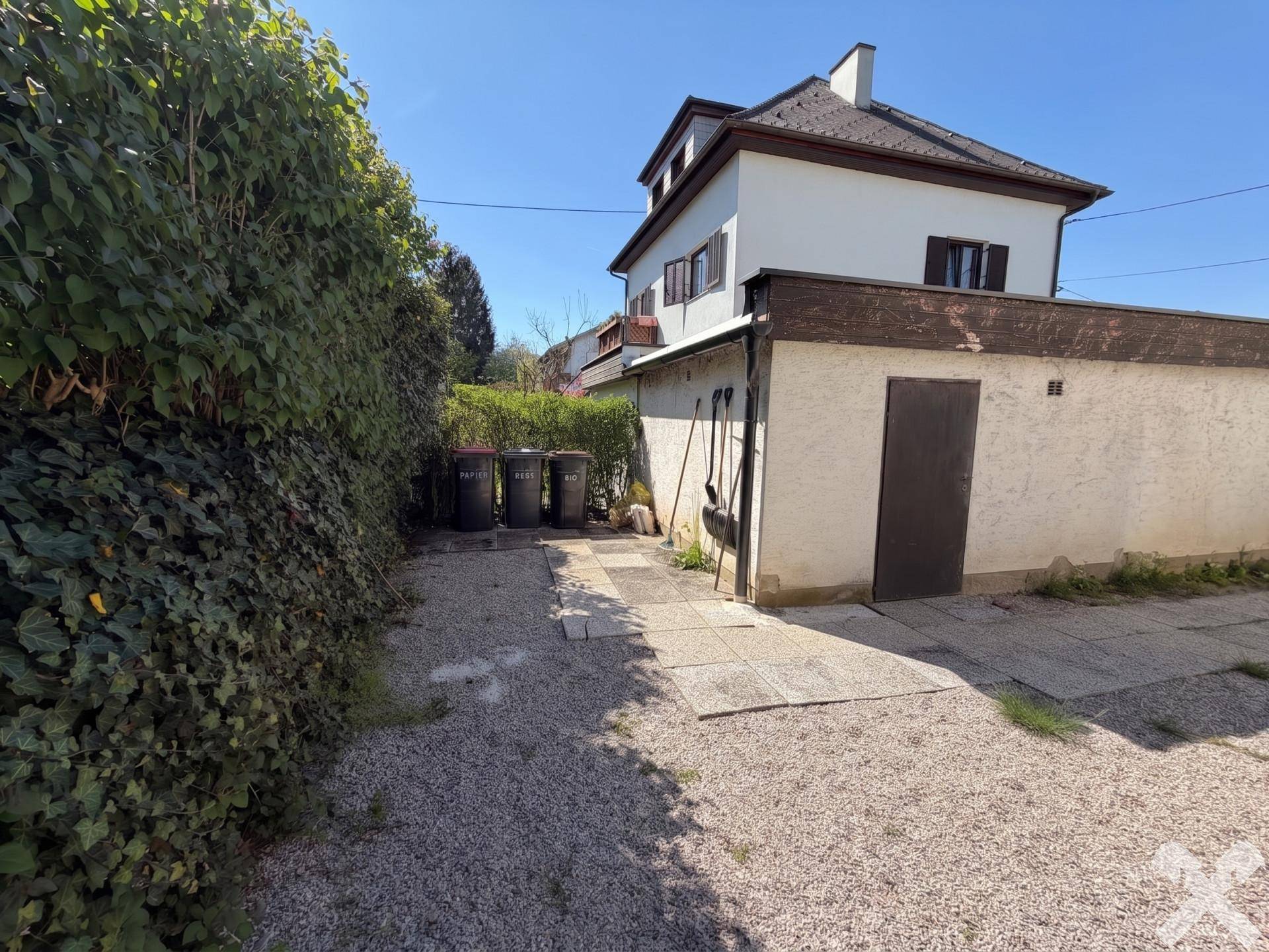 Seitenansicht des Hauses mit Nebengebäude, Kiesweg und Mülltonnen.