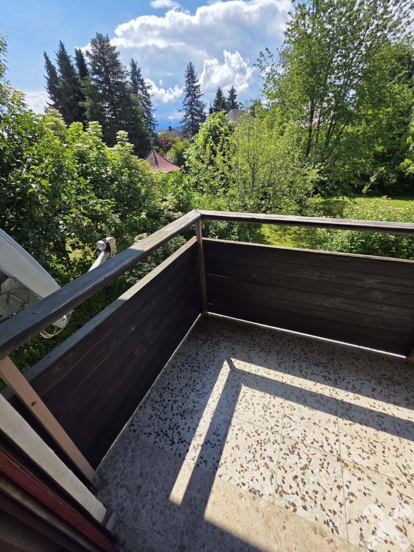 Eckbereich eines Balkons mit Fliesenboden und Holzgeländer, umgeben von Bäumen.