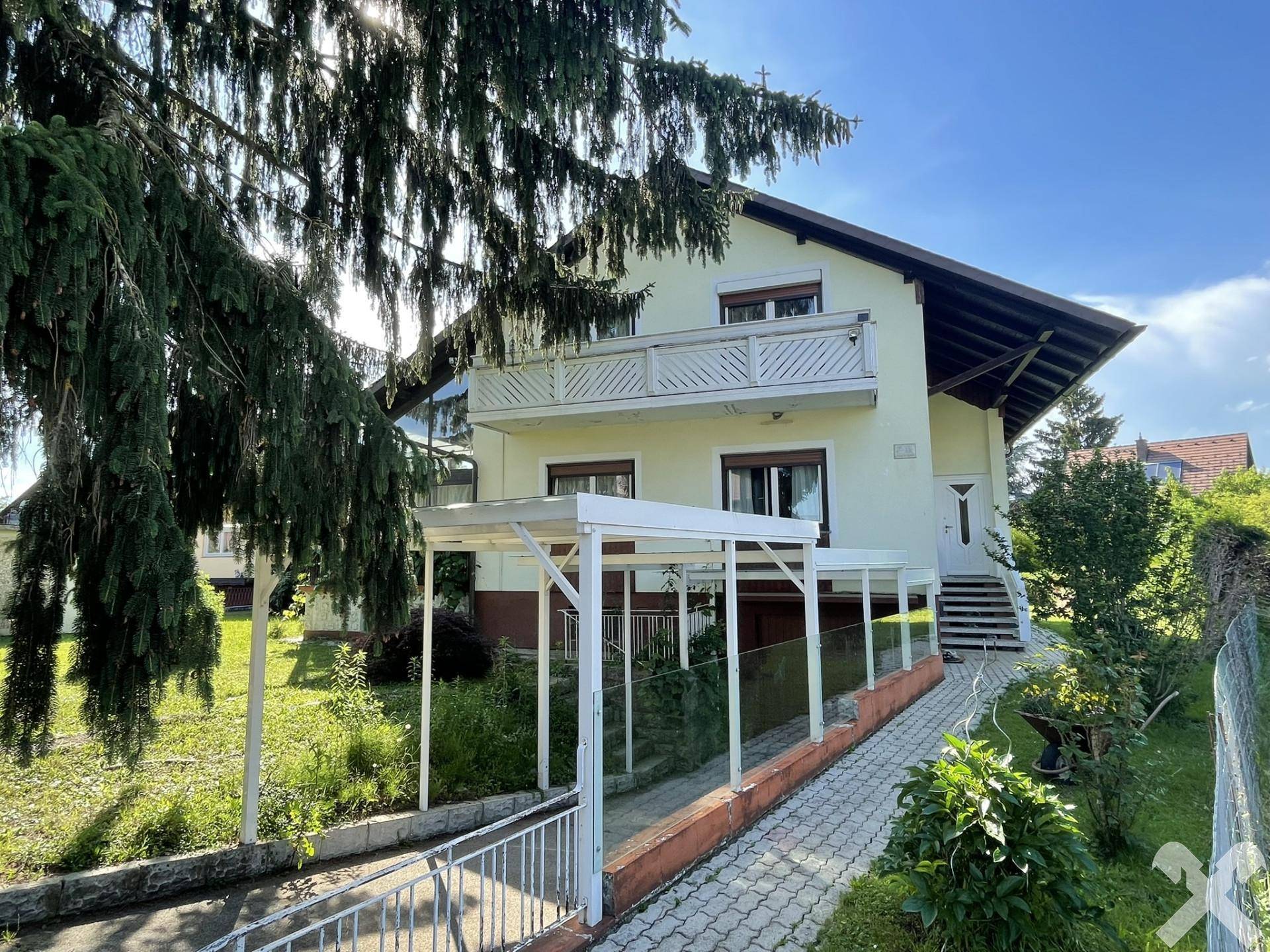 Vorderansicht des Hauses mit Balkon, Carport und gepflastertem Zugangsweg unter blauem Himmel.