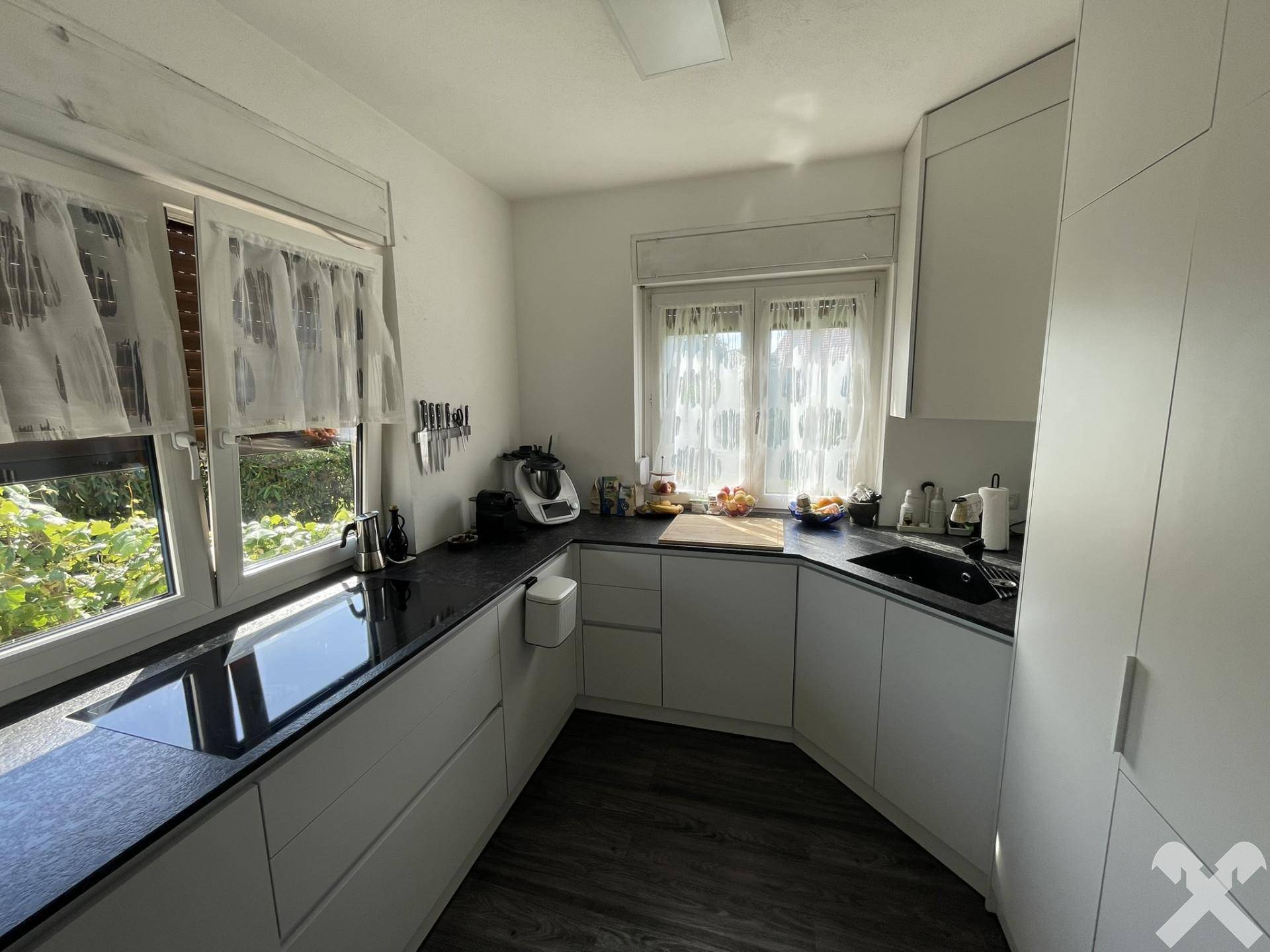 Moderne Küche mit weißen Schränken, dunkler Arbeitsplatte und Fensterblick ins Grüne.