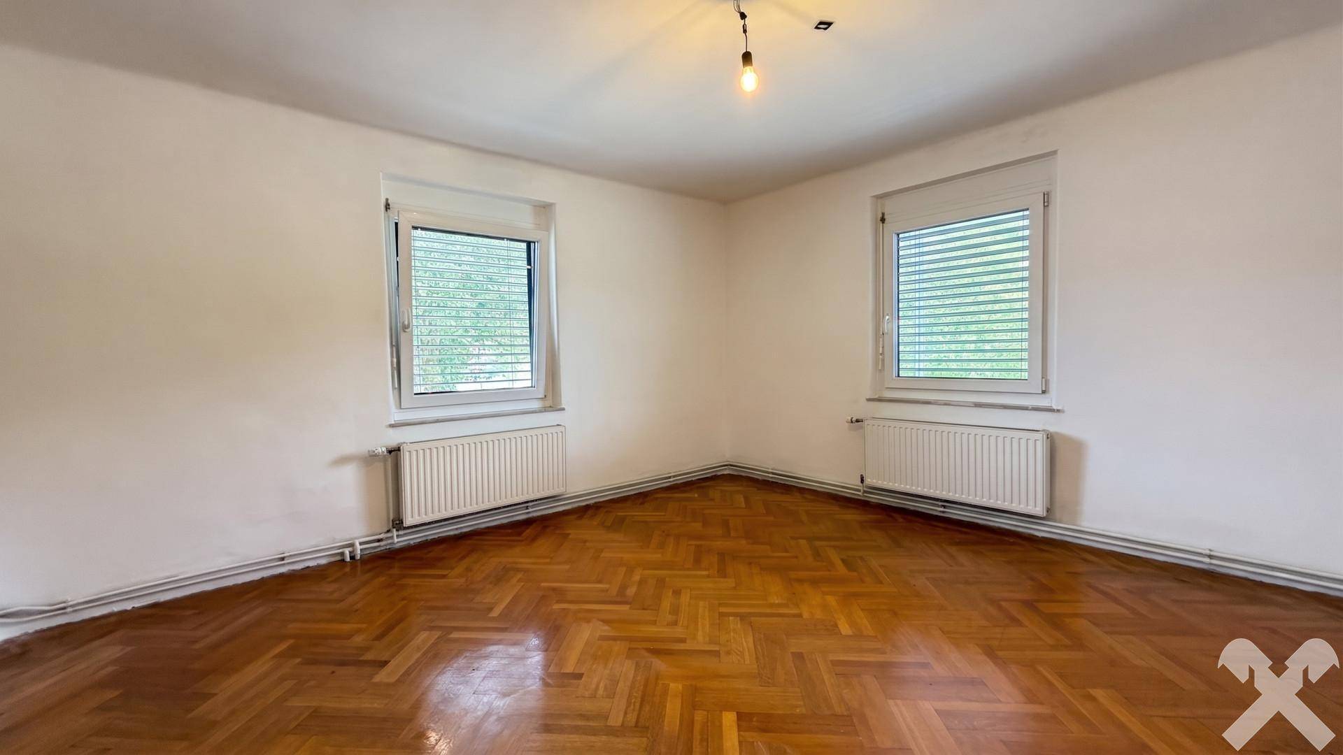 Helles, leeres Zimmer mit Fischgrätparkettboden und zwei Fenstern mit Heizkörpern.
