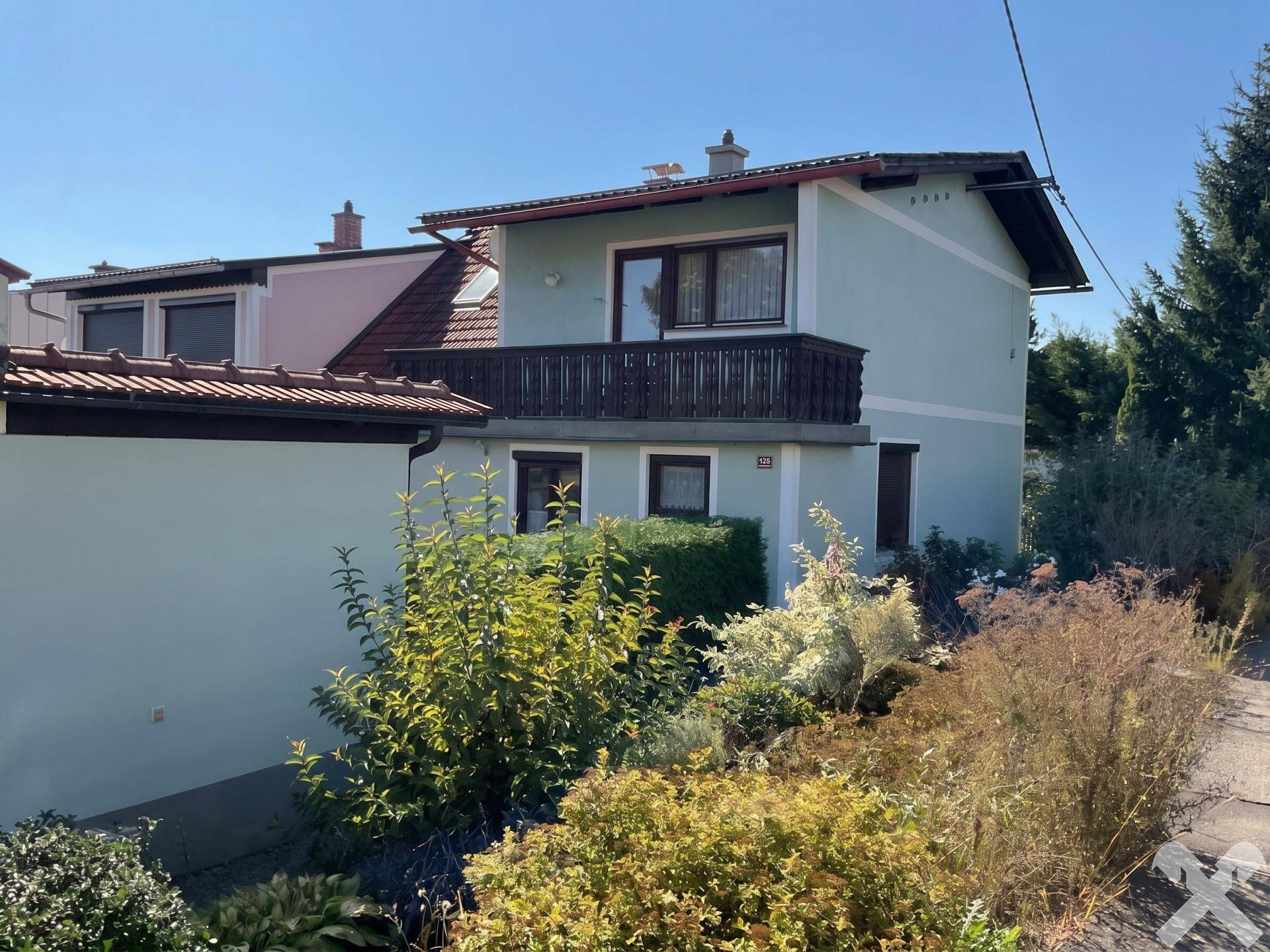 Seitenansicht des Hauses mit Balkon, grüner Fassade und üppiger Bepflanzung im Vorgarten.