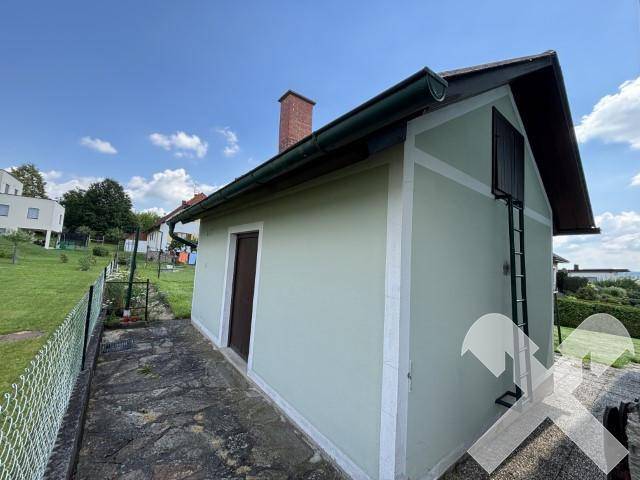 Seitenansicht des Hauses mit Anbau und umzäuntem Bereich unter blauem Himmel.