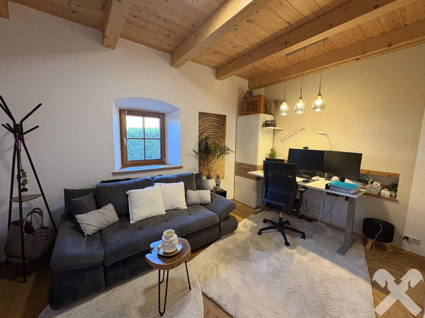 Helles Arbeitszimmer mit bequemer Couch, Schreibtisch und sichtbaren Holzbalken an der Decke.
