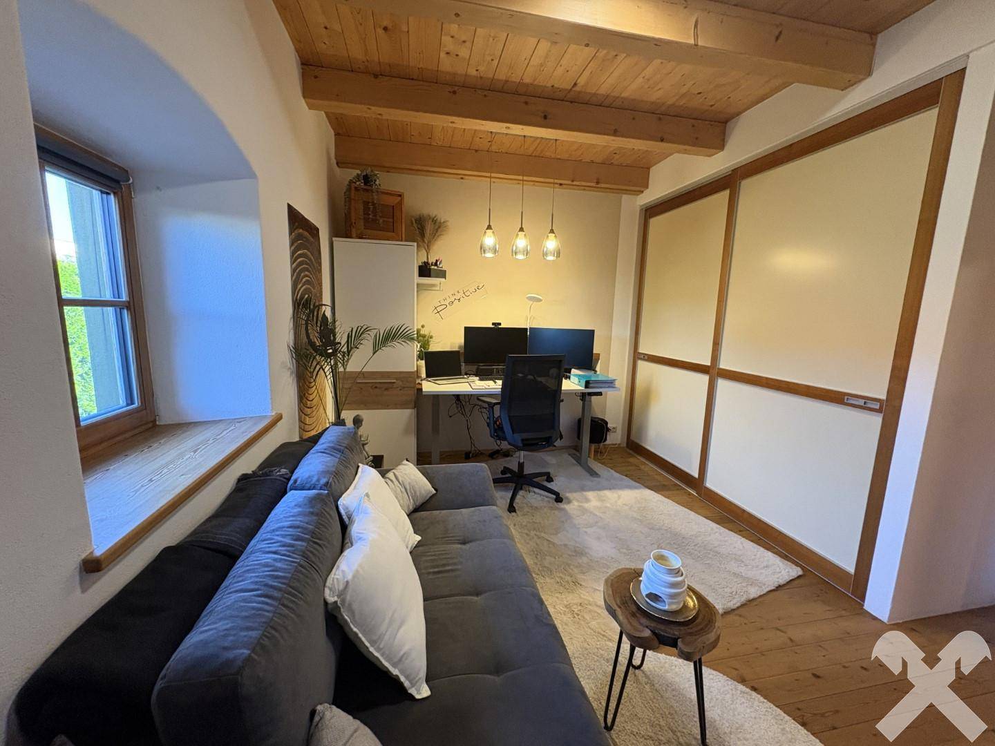 Geräumiges Büro mit viel Tageslicht, Sofa und integrierten Wandelementen unter der Holzbalkendecke.