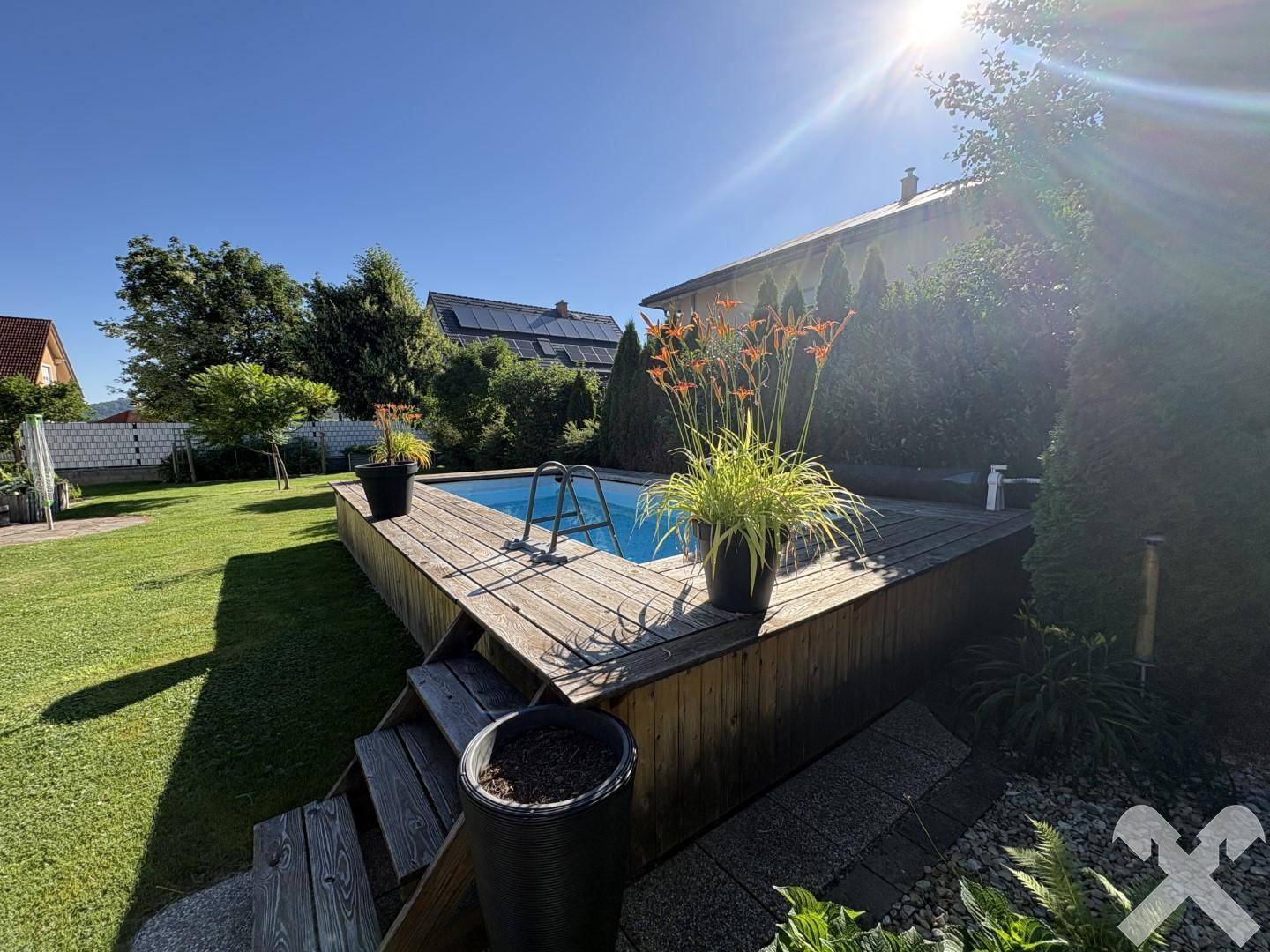 Sonnige Gartenanlage mit einem erhöhten Pool, Holzdeck und üppiger Bepflanzung.