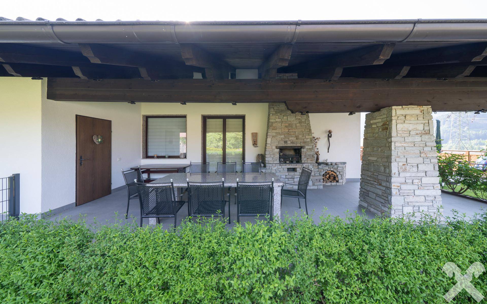 Überdachte Terrasse mit großem Esstisch, Steinkamin und Blick auf die grüne Berglandschaft.