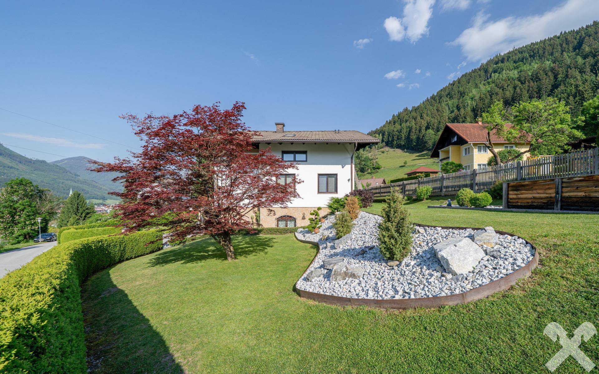 Gepflegter Gartenbereich mit Rasenfläche, Ziersträuchern und einem roten Ahornbaum vor bergiger Kulisse.