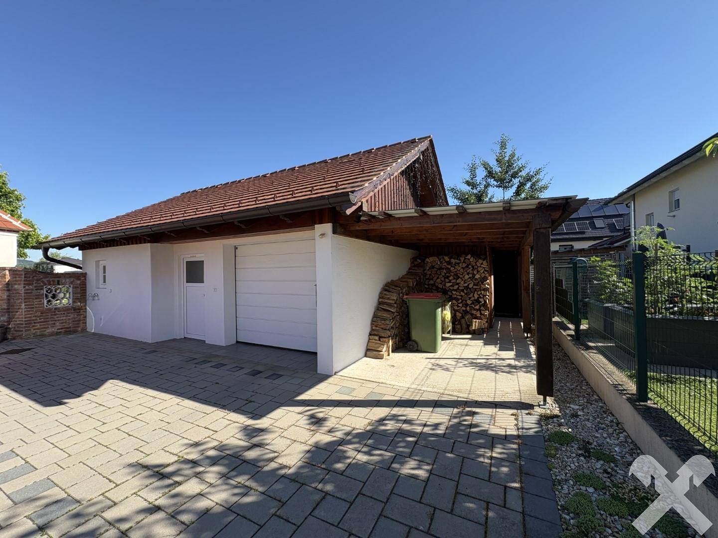 Garage mit Carport und Holzstapel, davor eine gepflasterte Einfahrt.