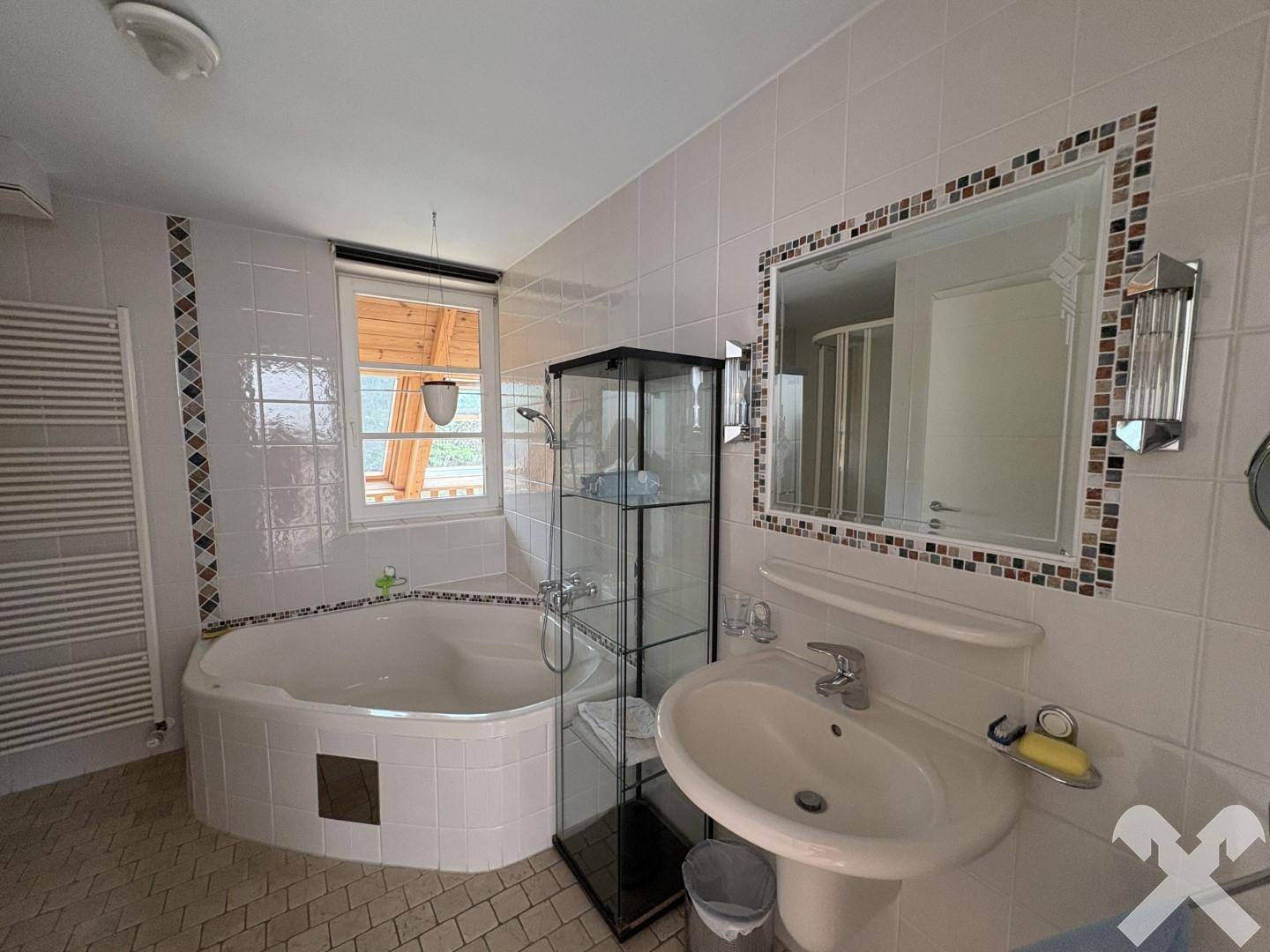 Badezimmer mit Eckbadewanne, Waschbecken, Fliesenboden und beheiztem Handtuchhalter.