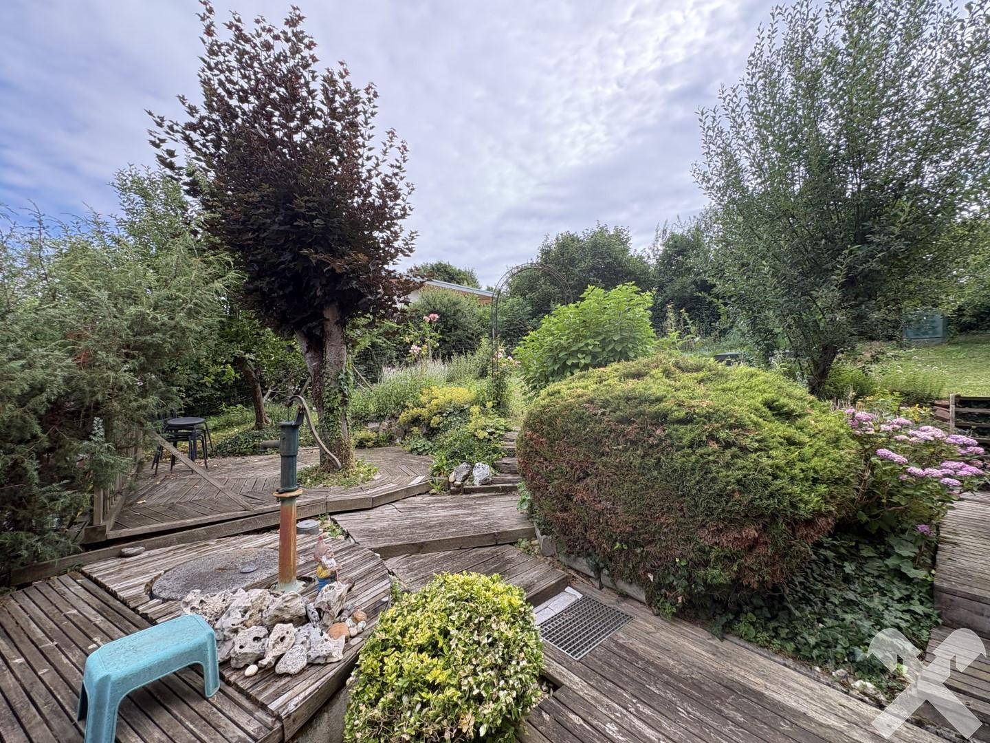 Terrassierter Garten mit Holzdecks, üppiger Bepflanzung und einer Wasserpumpe.