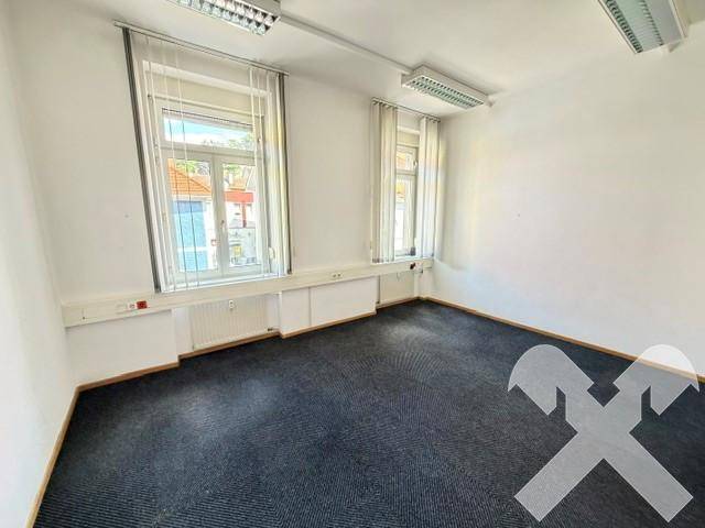 Kleinerer Büroraum mit Teppichboden, zwei Fenstern und Wandheizkörpern.