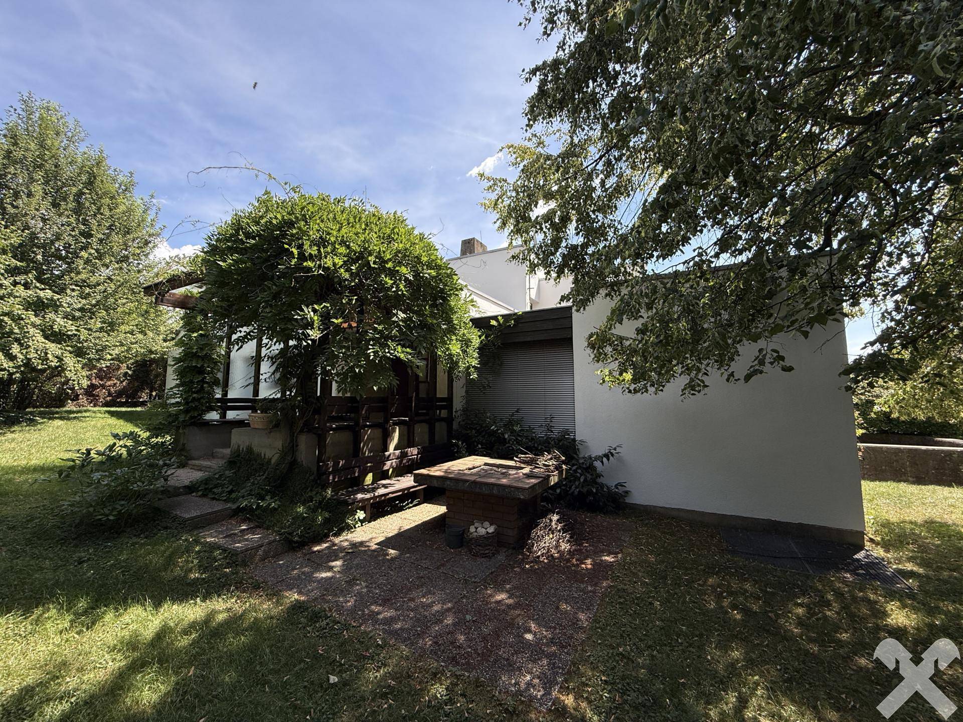 Grüner Gartenbereich mit üppiger Vegetation und einem kleinen gemauerten Grillplatz im Freien.