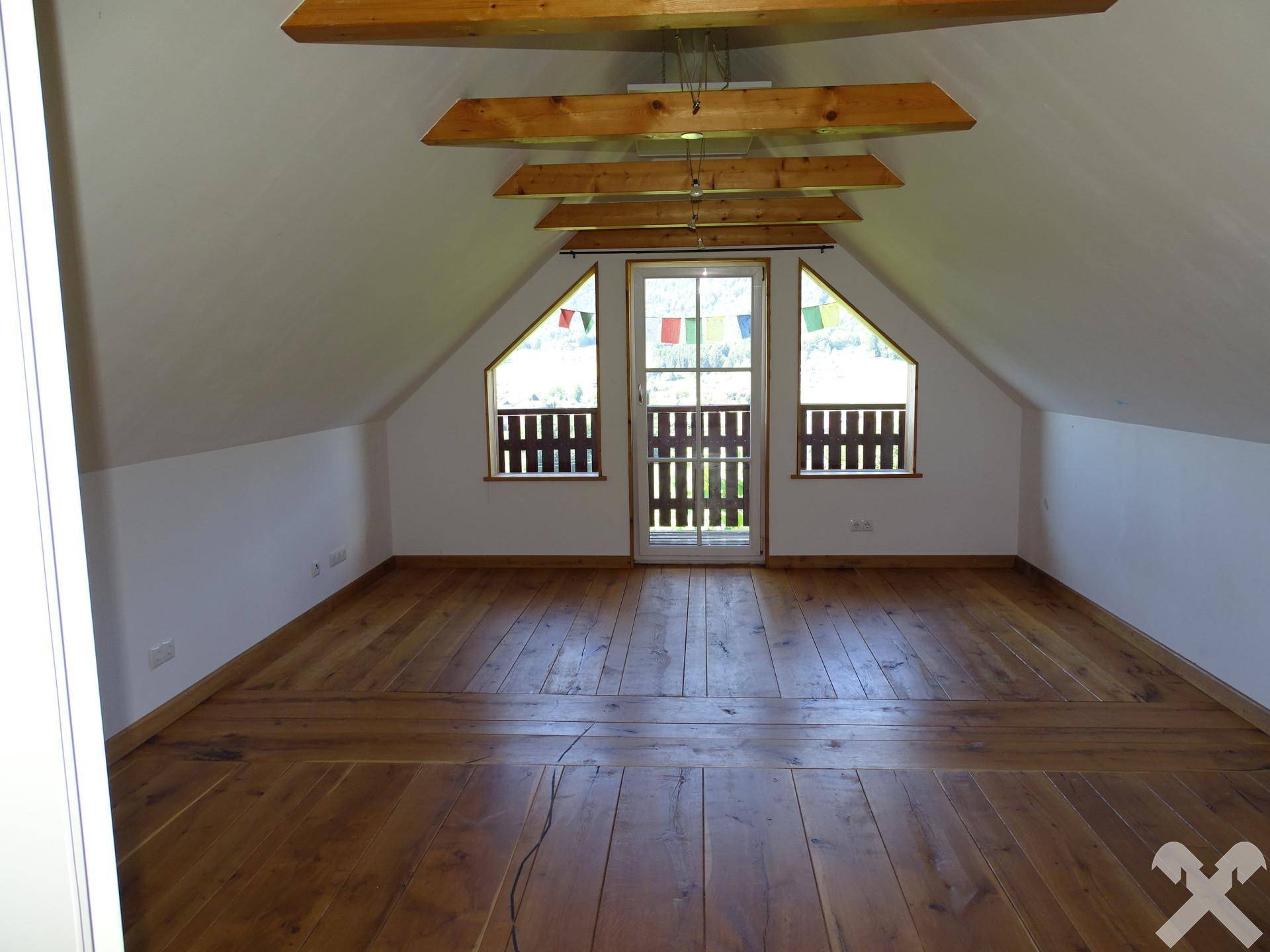Geräumiges Dachzimmer mit Holzboden, sichtbaren Holzbalken und Fenstern mit Blick ins Freie.