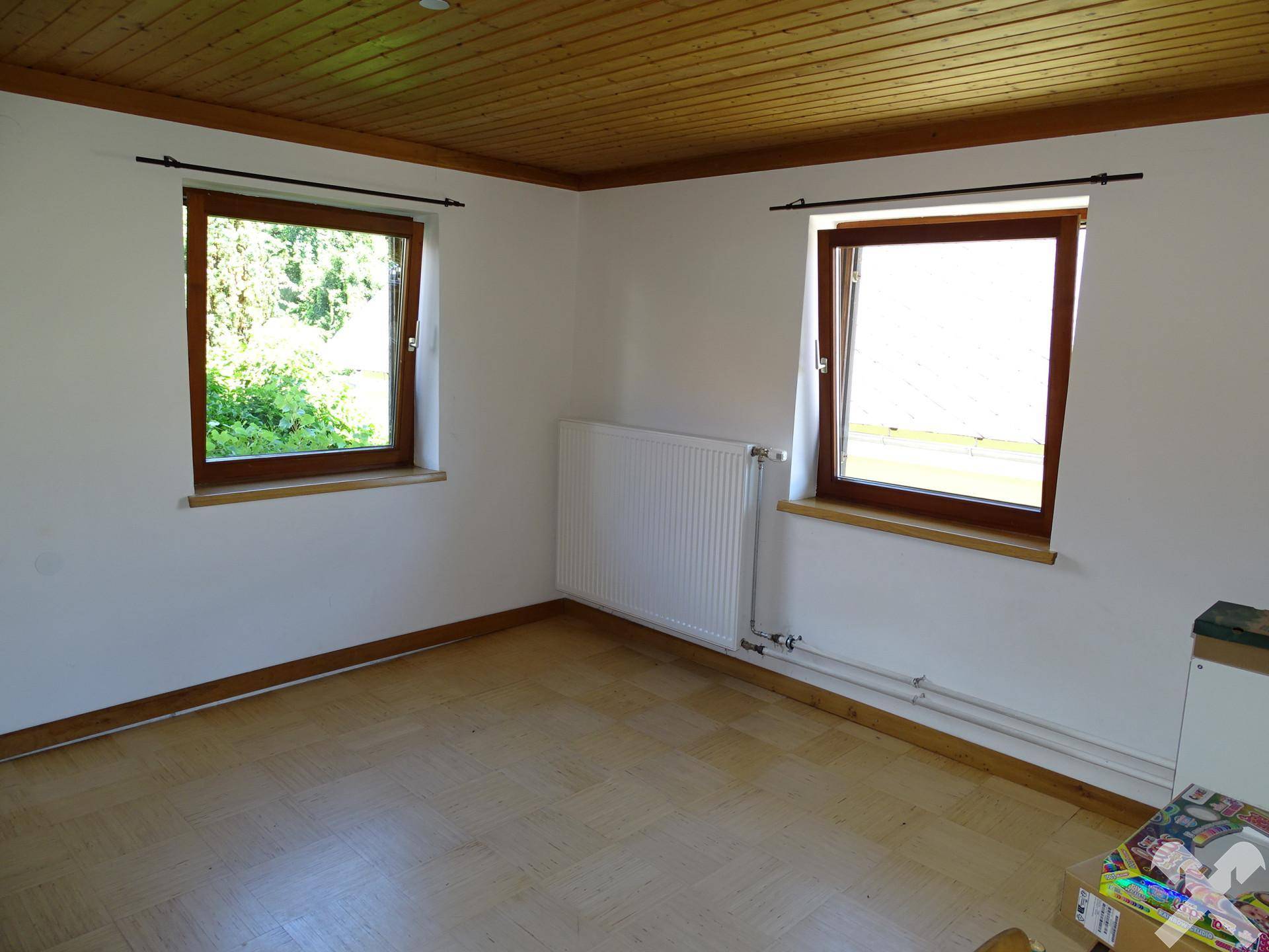 Helles Zimmer mit Holzdecke, Laminatboden und zwei Fenstern mit Blick ins Grüne.